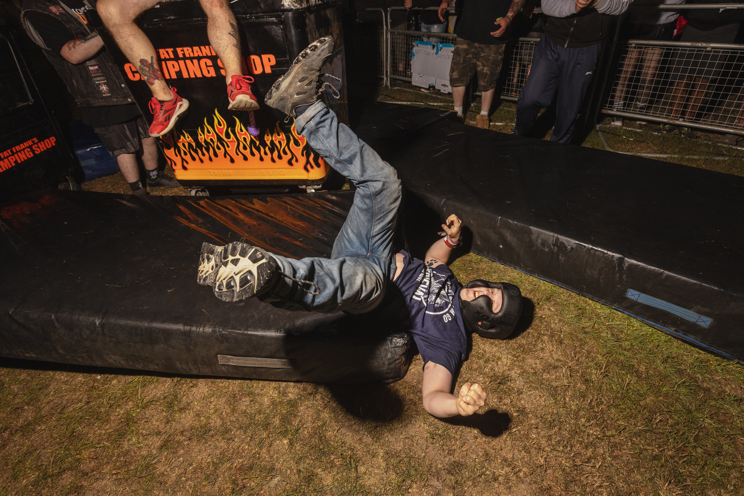 Person in denim jacket and spiked accessories lies on ground at outdoor event with crowd and barrier visible in background.