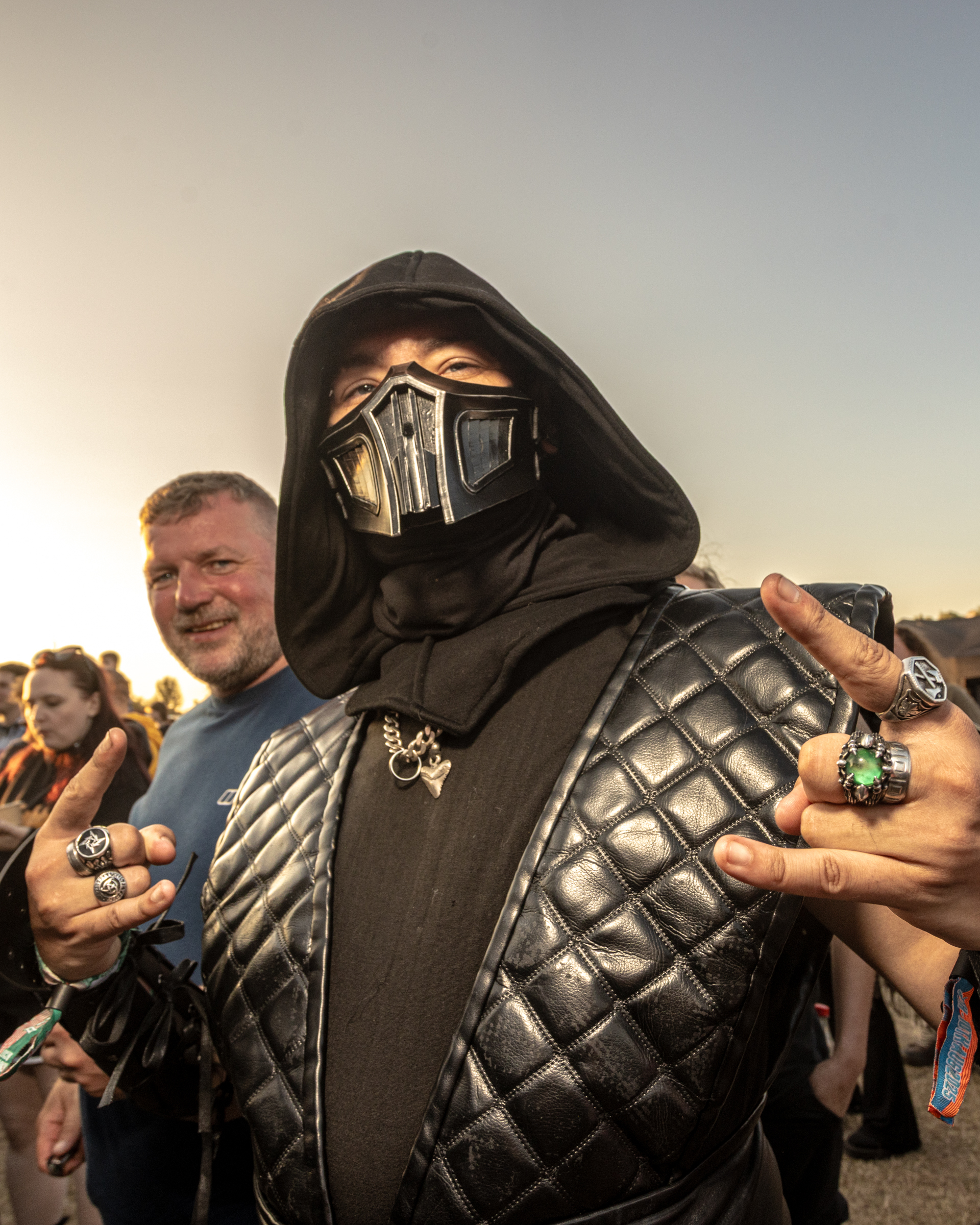 Person in black hooded costume with metallic mask and quilted armour at outdoor event, surrounded by crowd reaching towards them.