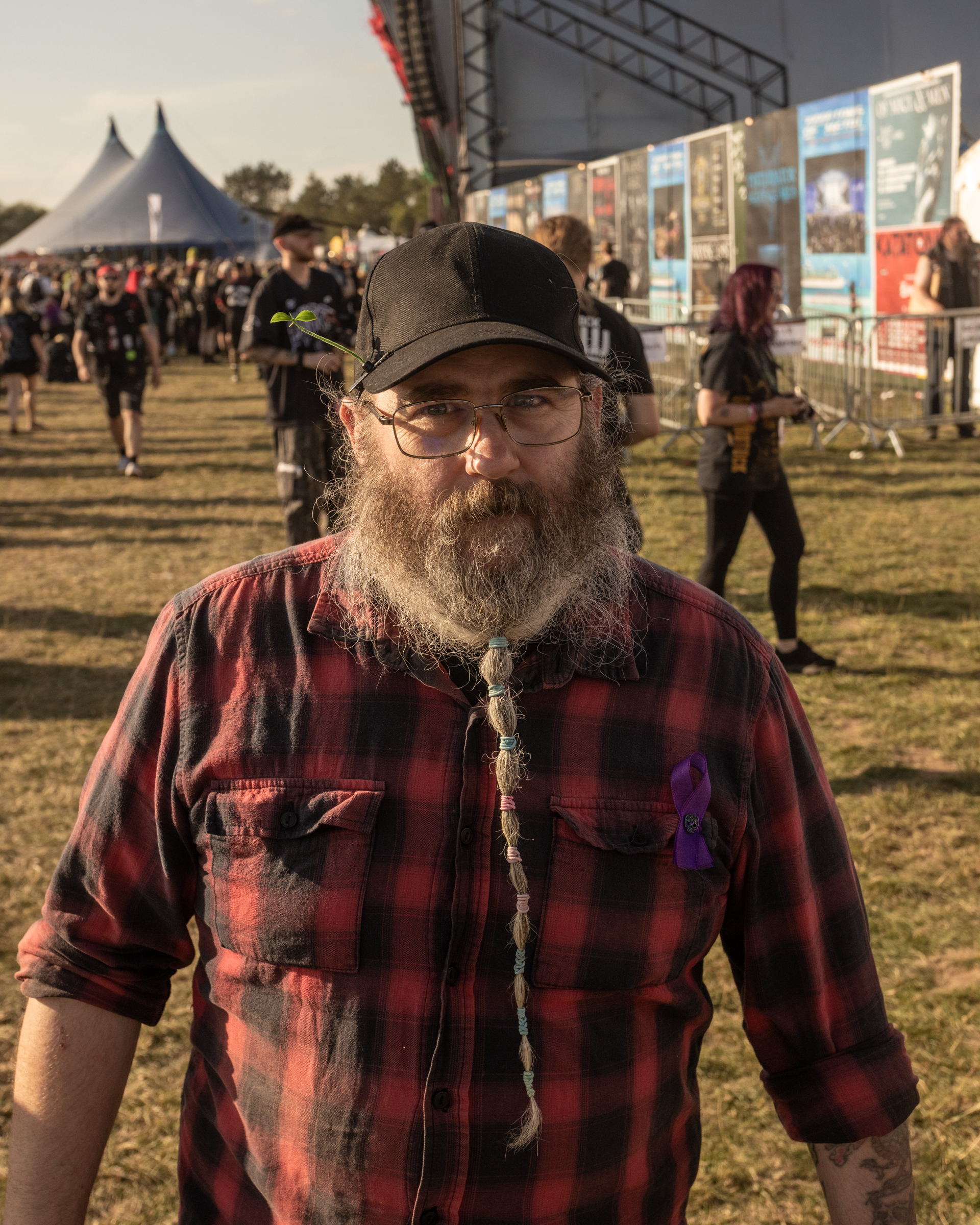 Bearded man in glasses, cap, and red plaid shirt at outdoor festival with stage tents and crowd in background.