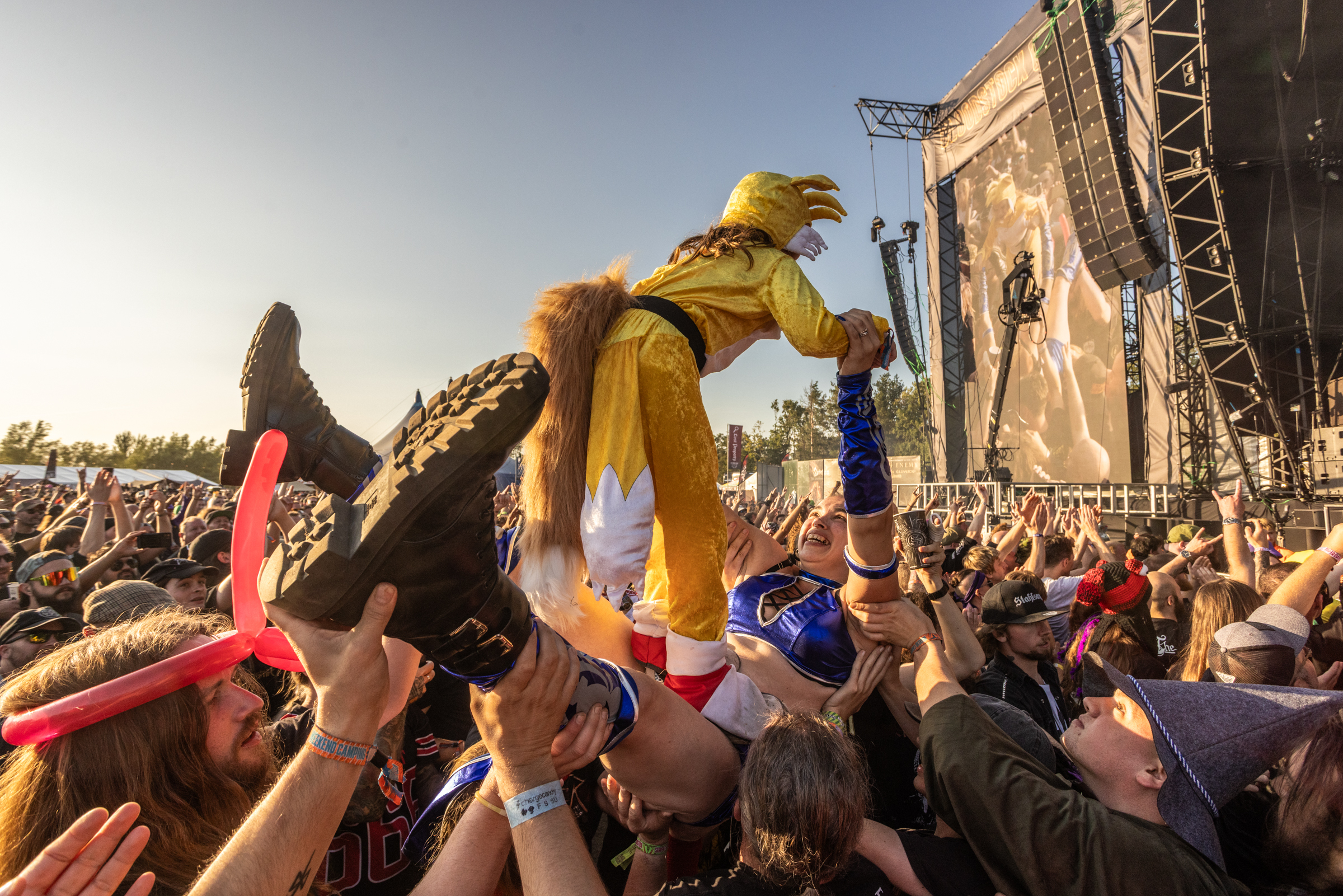 Person in yellow outfit crowd-surfing over festival audience with hands reaching up, stage and metal barriers visible in background.
