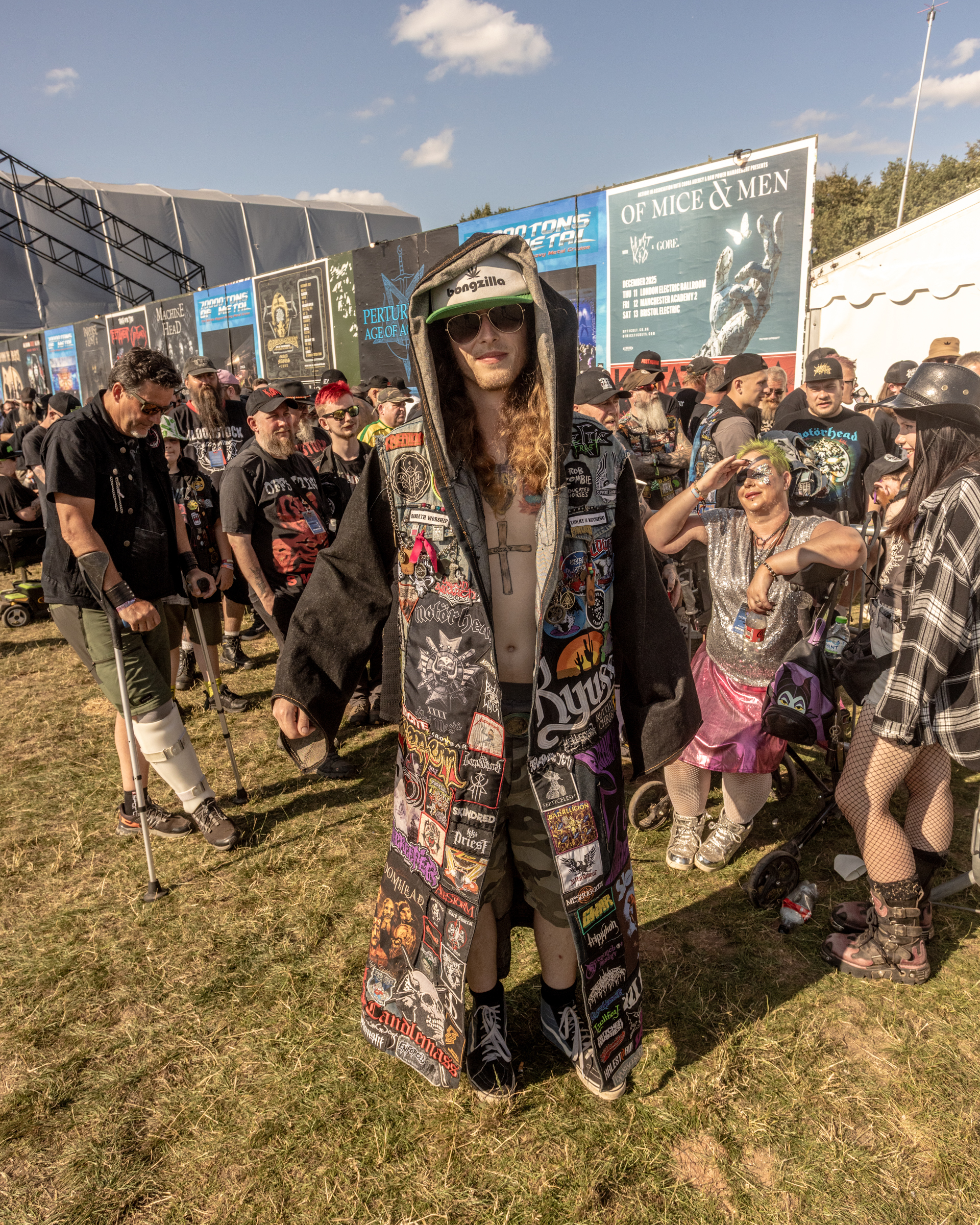Woman in hooded coat covered with band patches stands amongst crowd at outdoor music festival with blue signage visible.