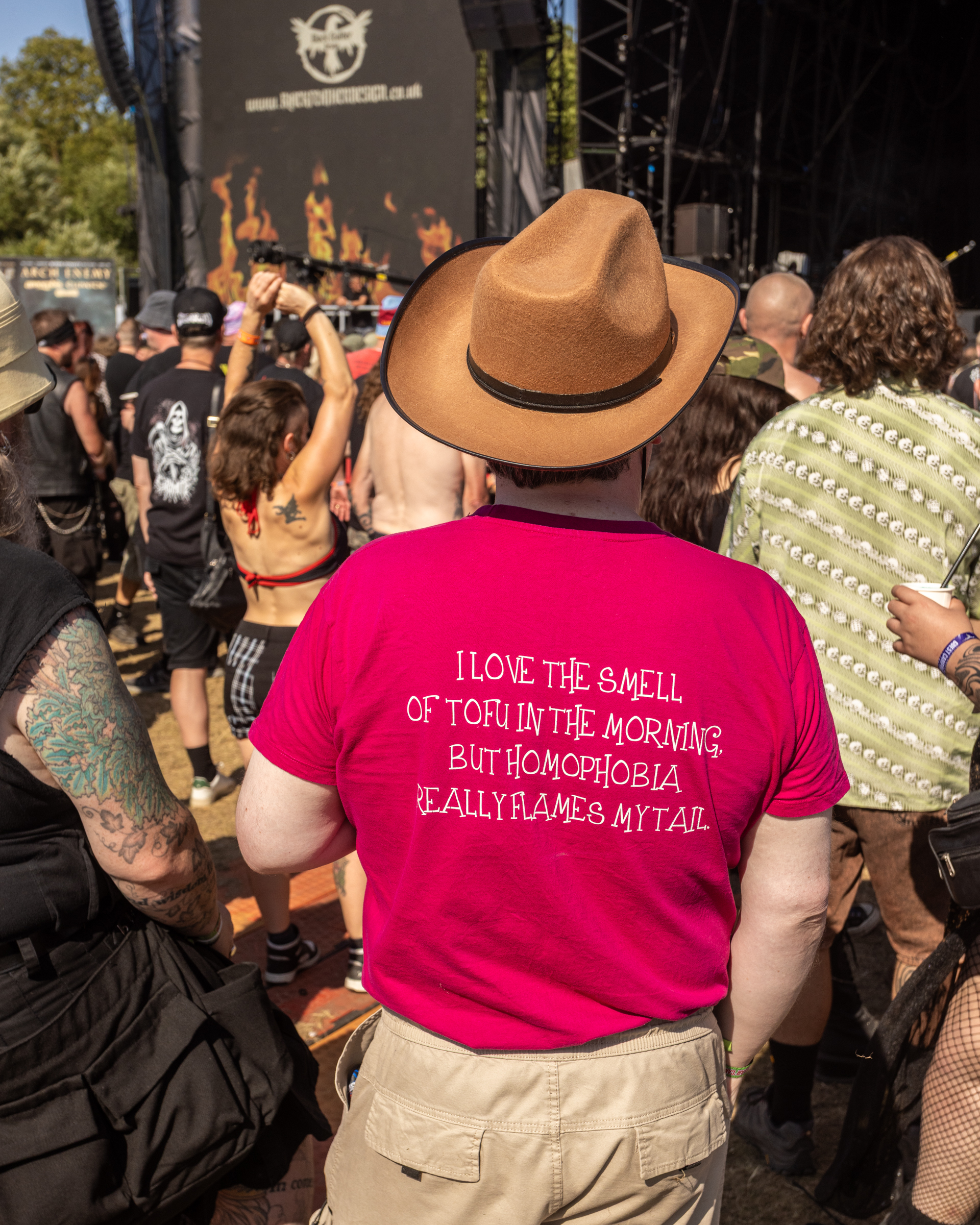Person in brown cowboy hat and pink t-shirt with humorous text about tofu and homophobia at outdoor music festival crowd.