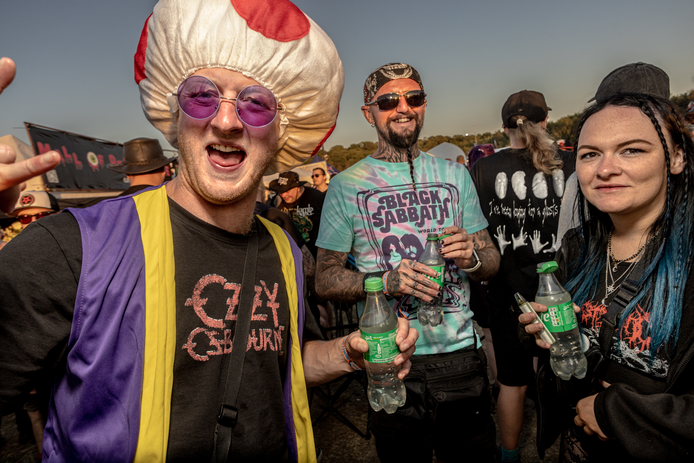 Three people in costume at outdoor event - person in blonde turban and purple glasses, bearded man in blue t-shirt, woman with blue hair.