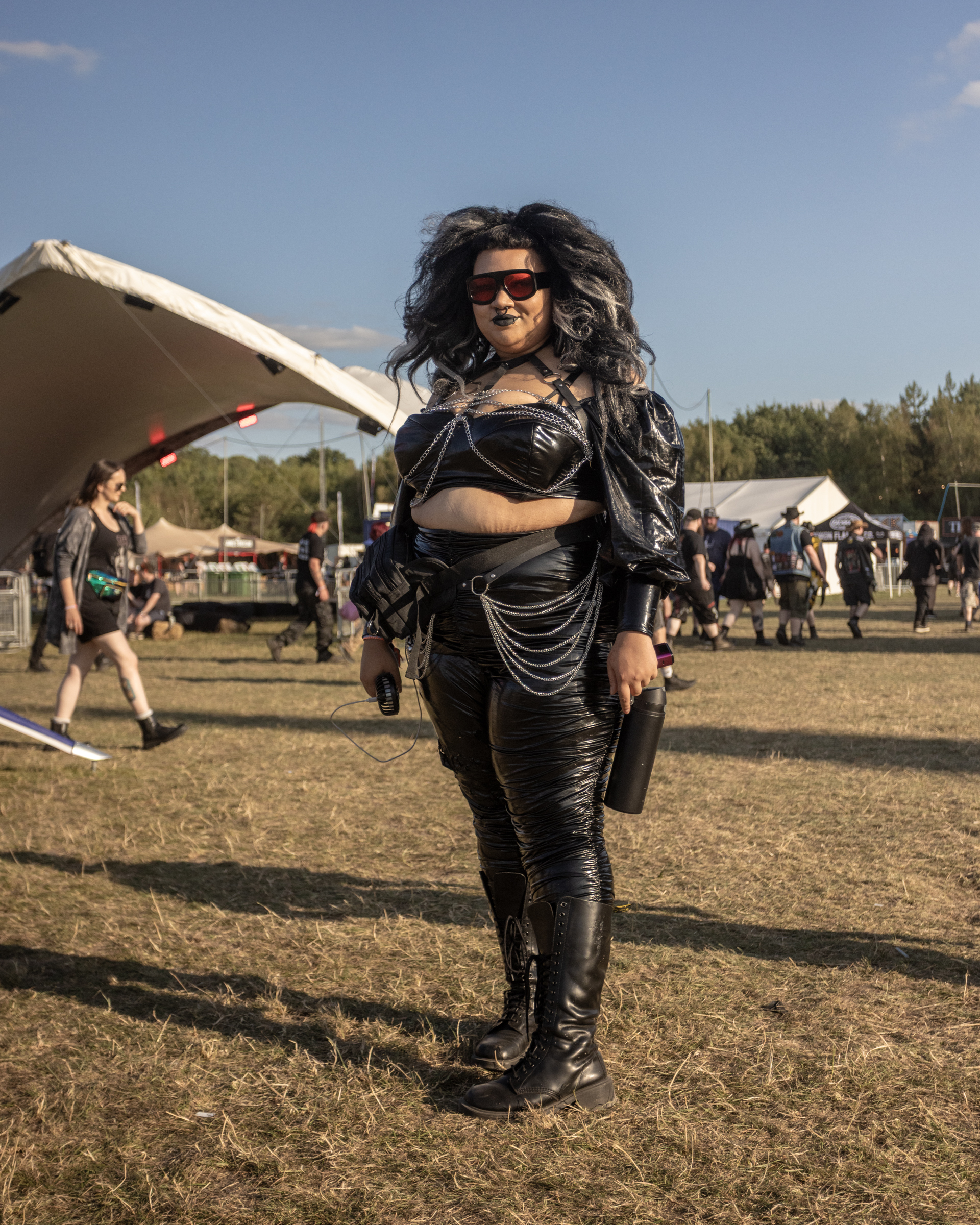 Woman in black leather outfit with sunglasses and curly hair at outdoor festival, tent and crowd in background.