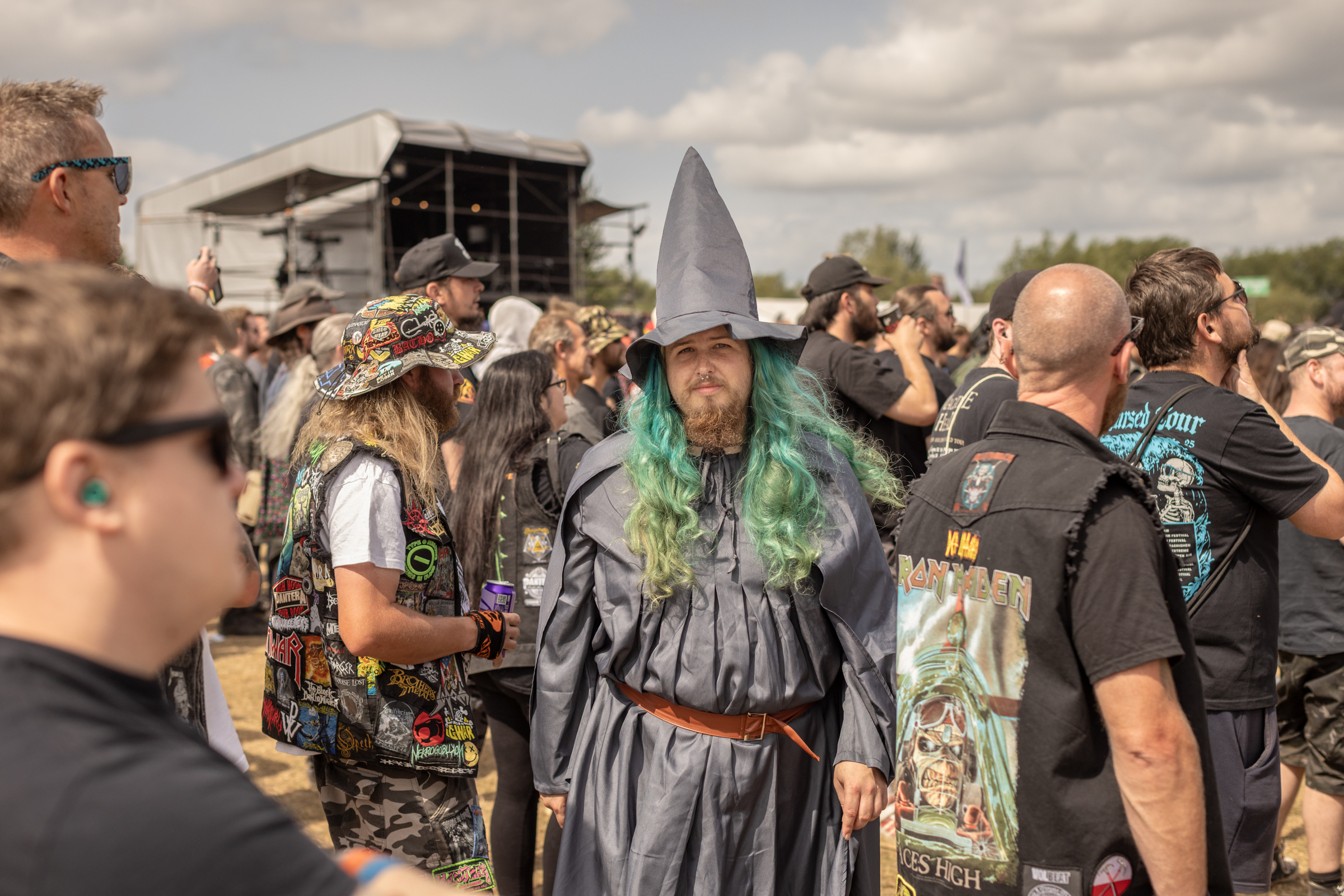 Man in grey wizard costume with pointed hat and green garland amongst crowd at outdoor event with stalls and cloudy sky.