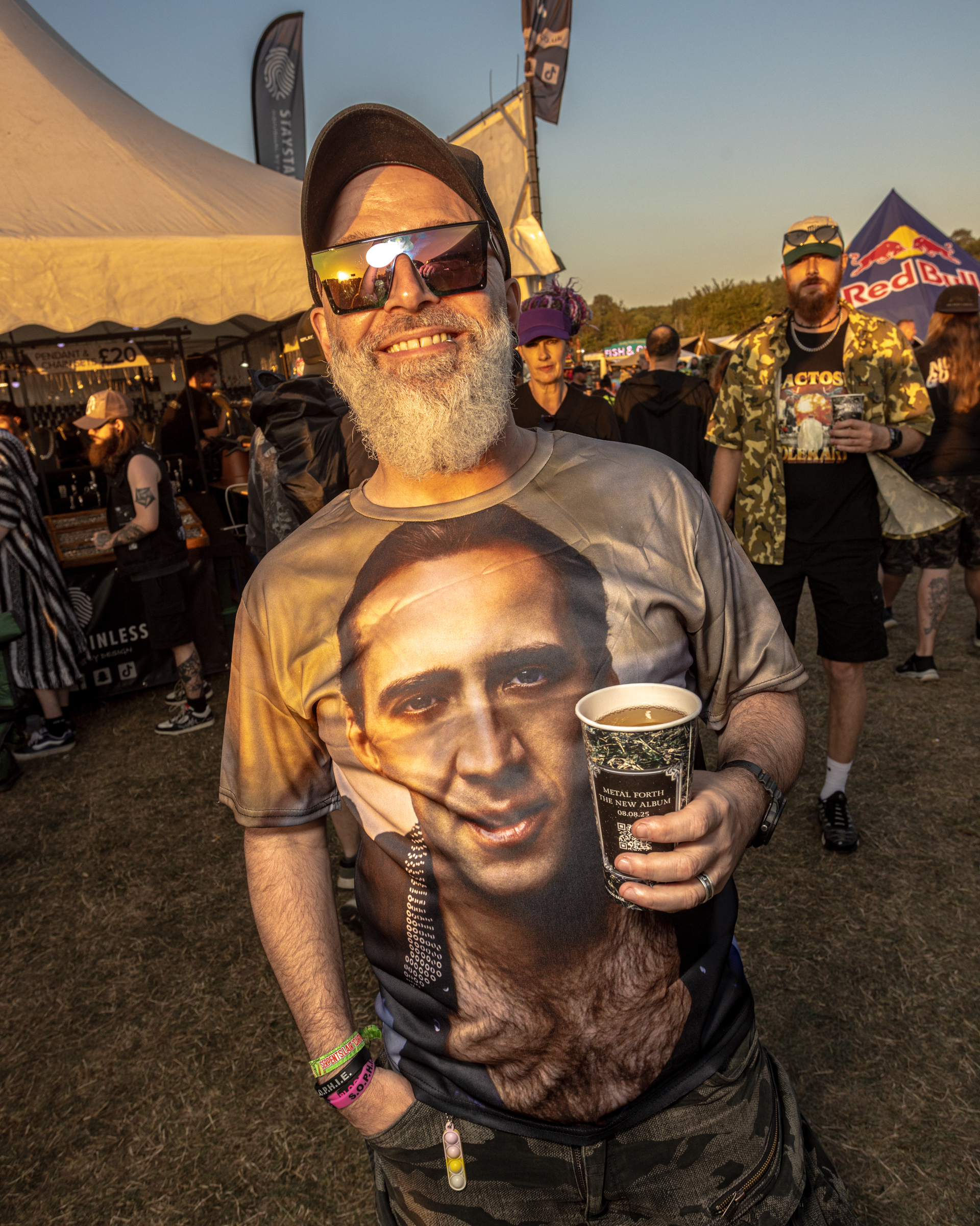 Bearded man in sunglasses wearing Nicolas Cage t-shirt holds coffee cup at outdoor festival with tents and crowds in background.