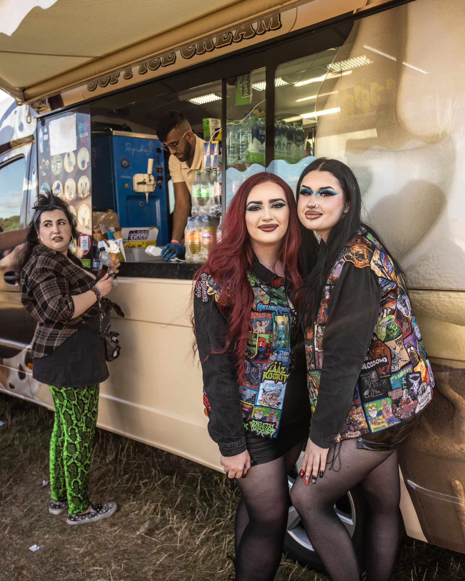 Three people in alternative fashion at outdoor food stall. Two women pose together wearing colourful patch jackets and dark makeup.