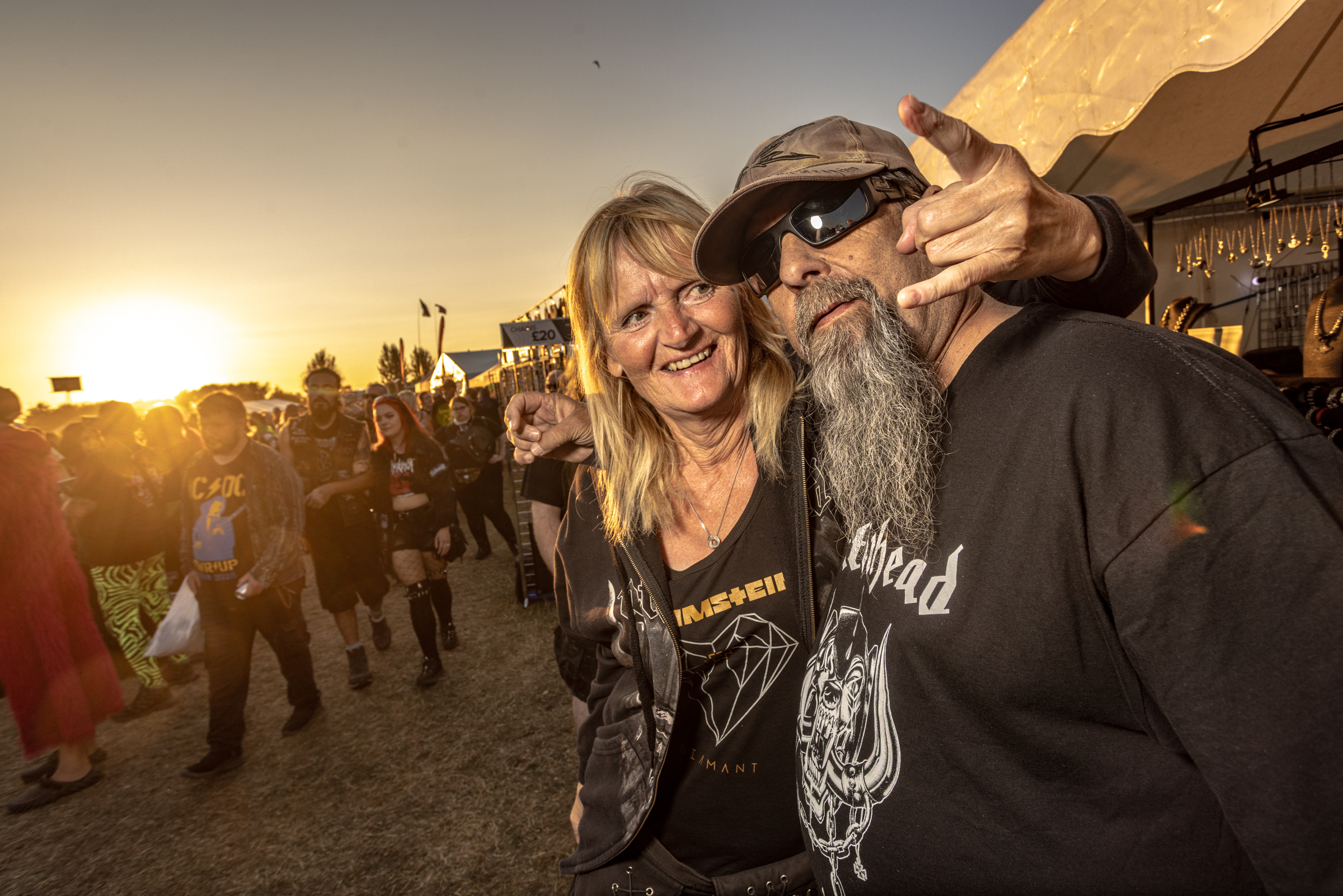 Two people making peace signs at outdoor event during golden hour sunset, man with long grey beard wearing sunglasses and cap.