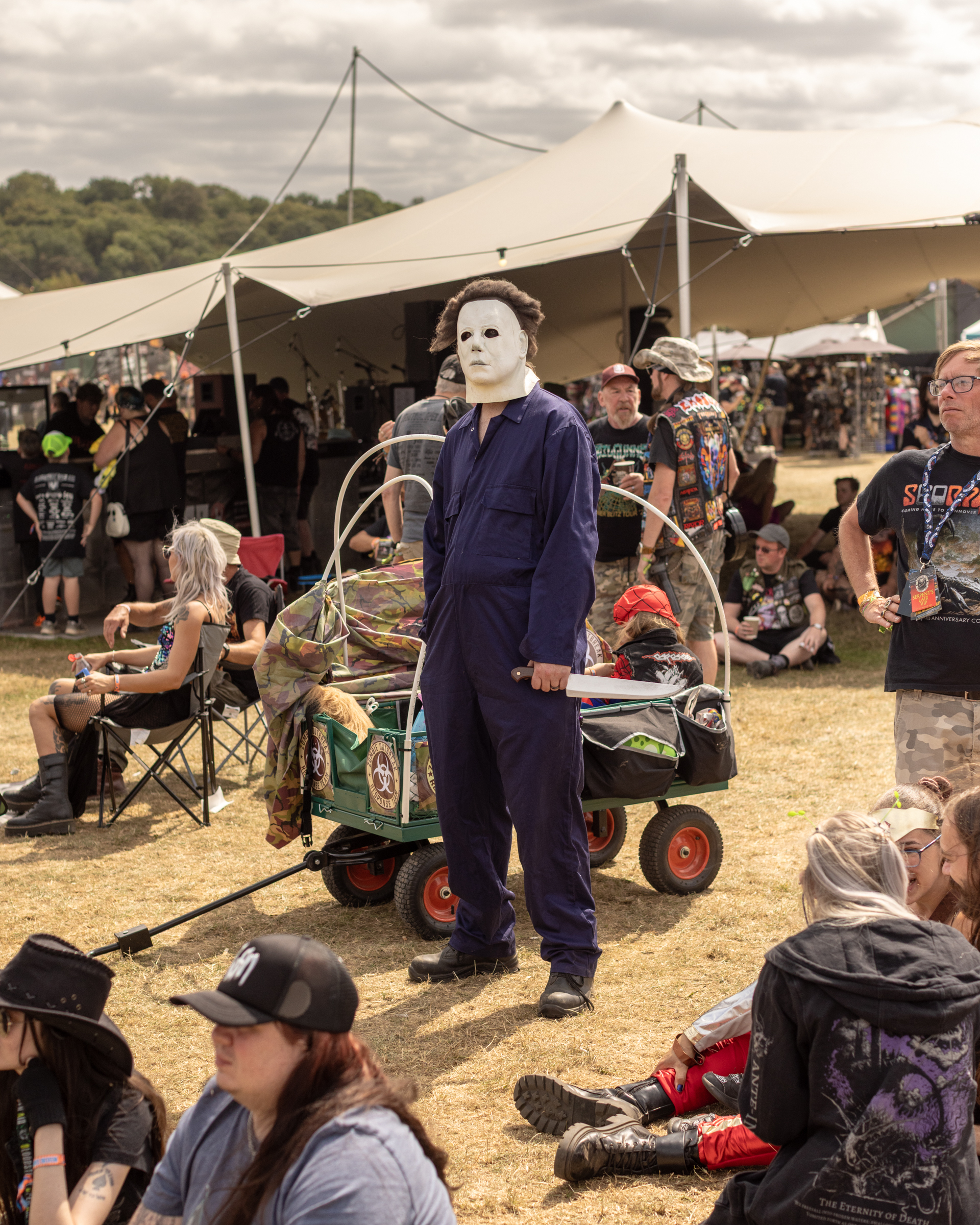 Person in white mask and dark blue boilersuit stands beside decorated cart at outdoor event with white marquee and crowd seated on grass.