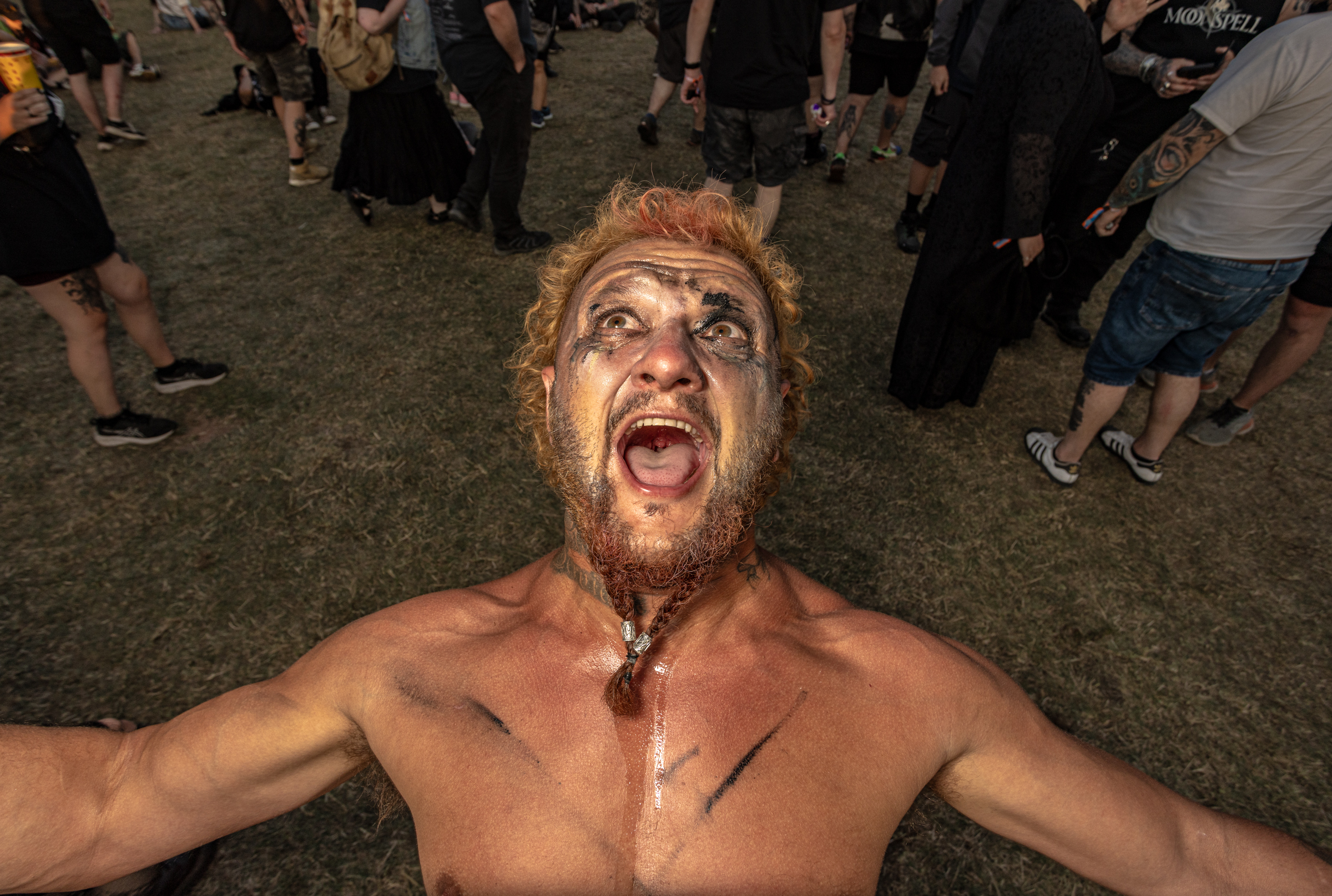 Shirtless man with blonde hair and face paint screaming whilst surrounded by crowd at outdoor event.