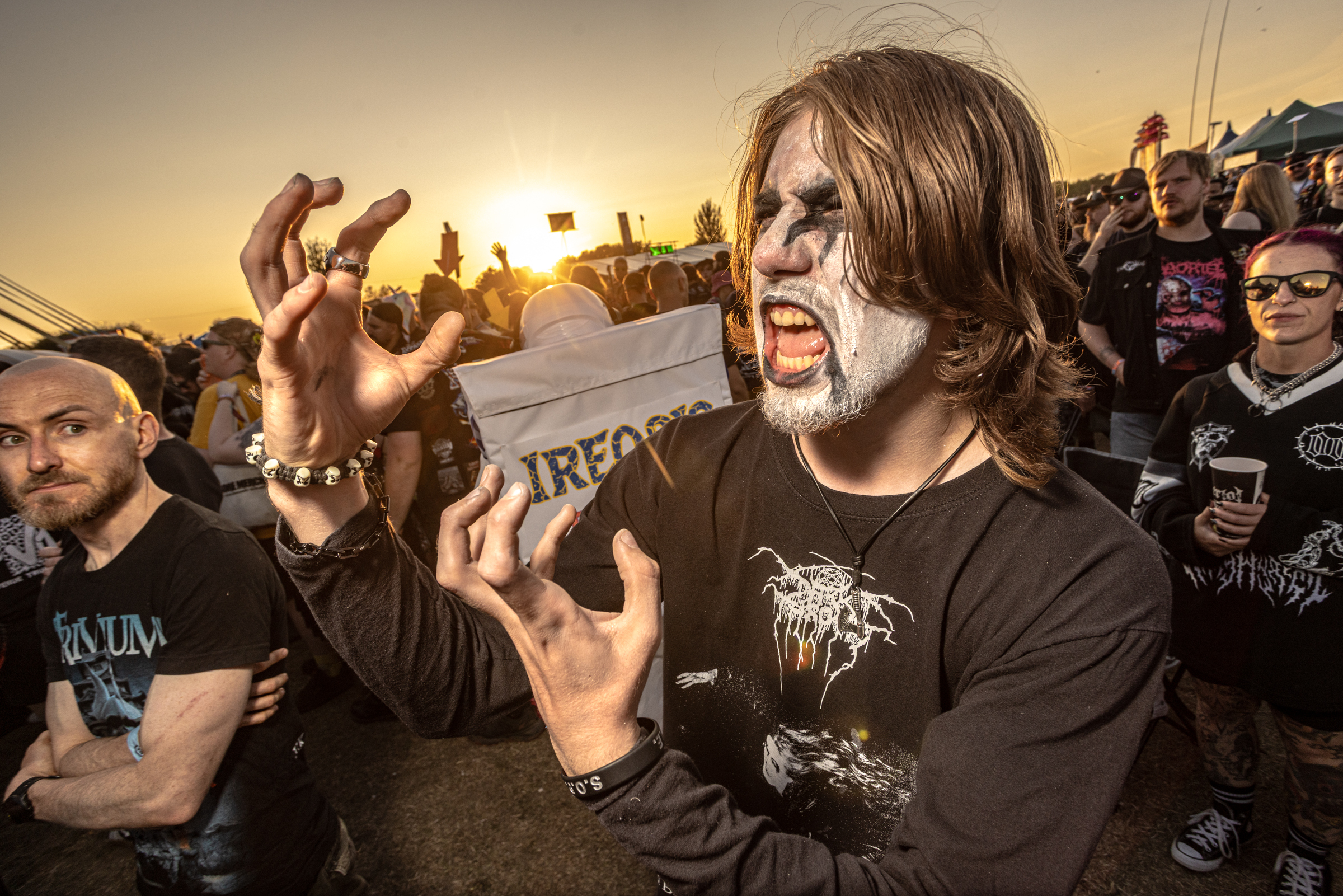 Long-haired person in black metal band shirt making aggressive gesture with raised hands amidst crowd at outdoor concert, sunset sky.