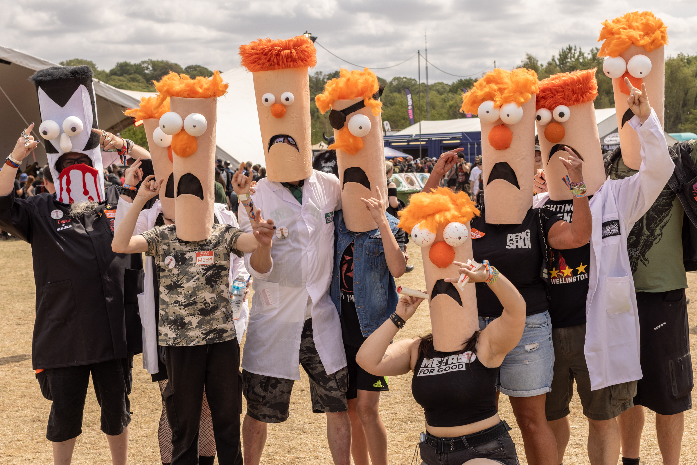 Group of people holding orange puppet heads with googly eyes and black moustaches at outdoor event, cloudy sky background.