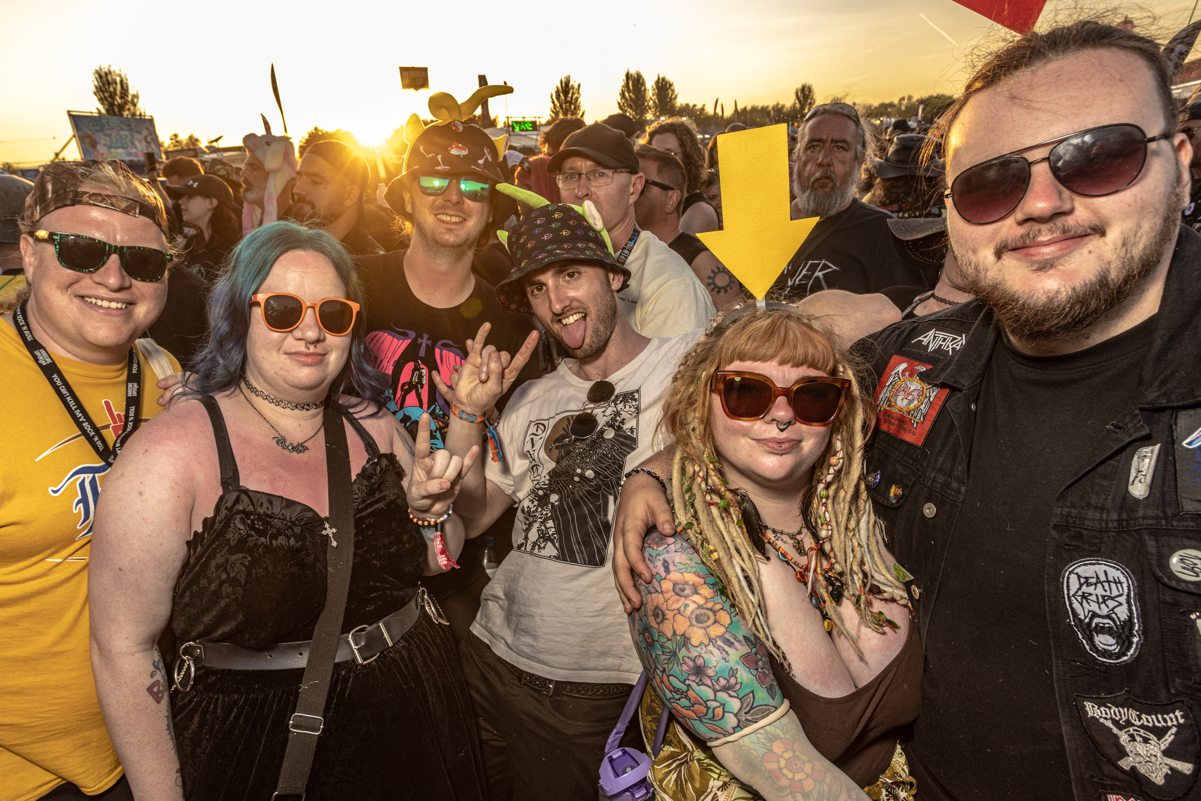 Group of seven people in sunglasses and festival attire posing together at outdoor event with bright yellow sky and crowd in background.