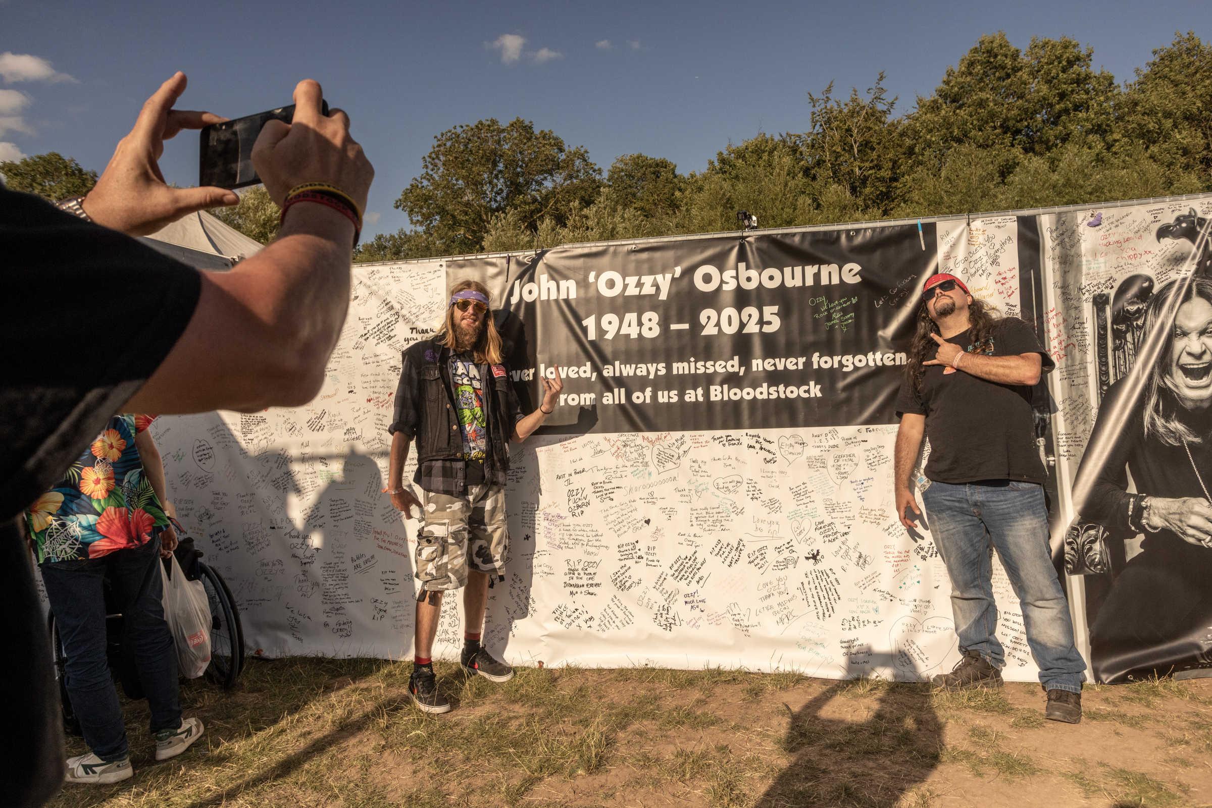People gathering at concrete memorial wall with John "Ozzy" Osbourne 1948-2025 tribute banner, person filming with mobile phone.