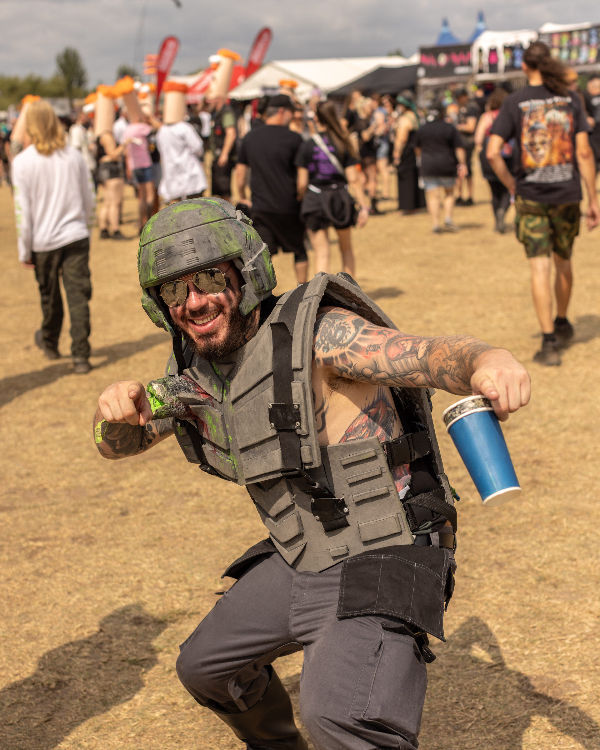 Tattooed man in military gear and helmet crouches at outdoor festival, holding blue drink can, with crowd and stalls behind