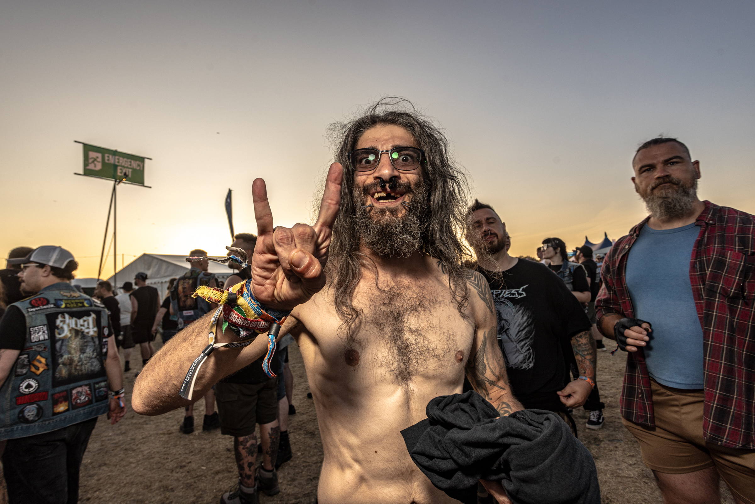 Shirtless bearded man with sunglasses making peace signs at outdoor festival, crowd and tents visible in background at sunset.