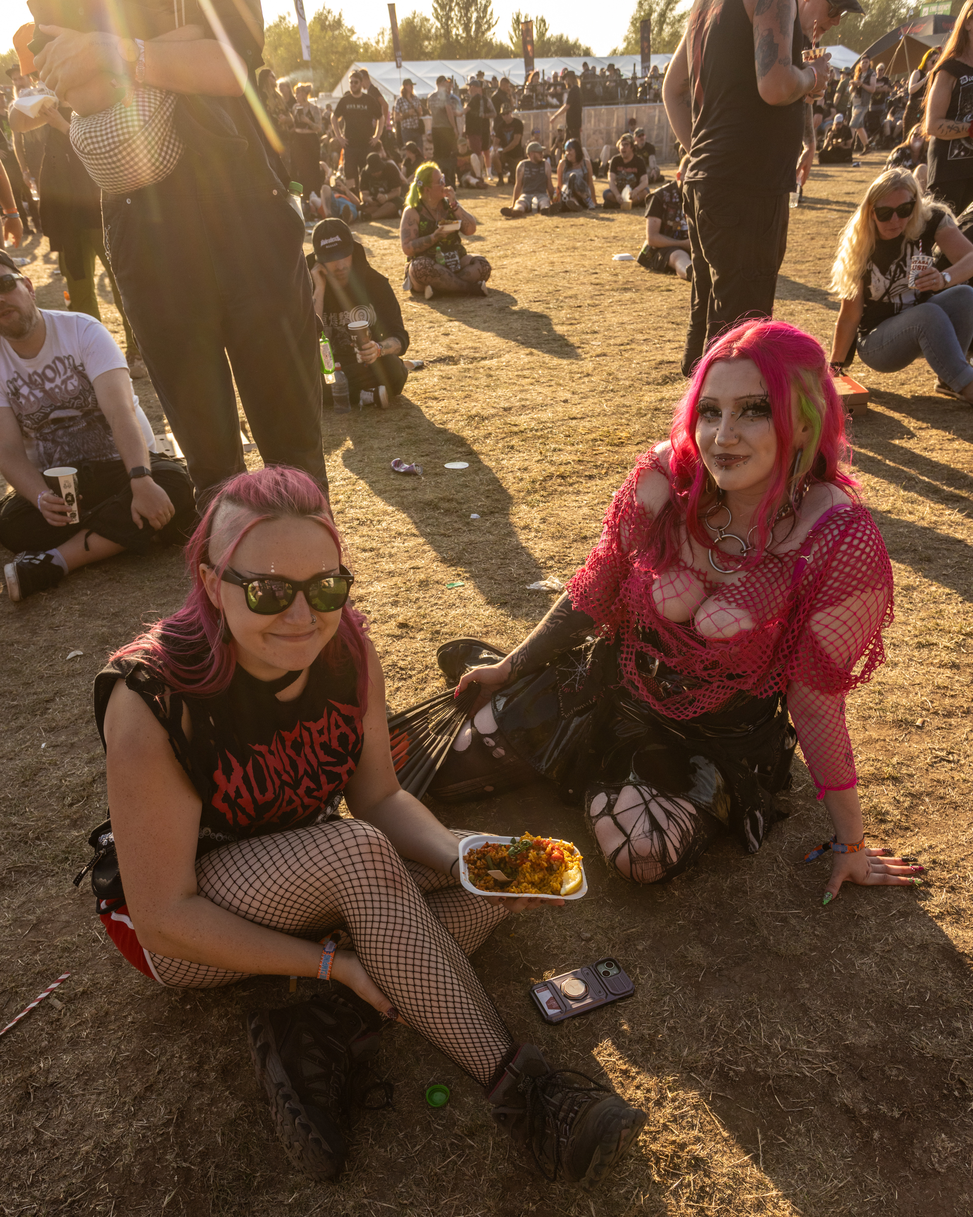 Two women sitting on dusty ground at outdoor festival, one in fishnet tights and sunglasses, other in bright pink headscarf, crowd in background.