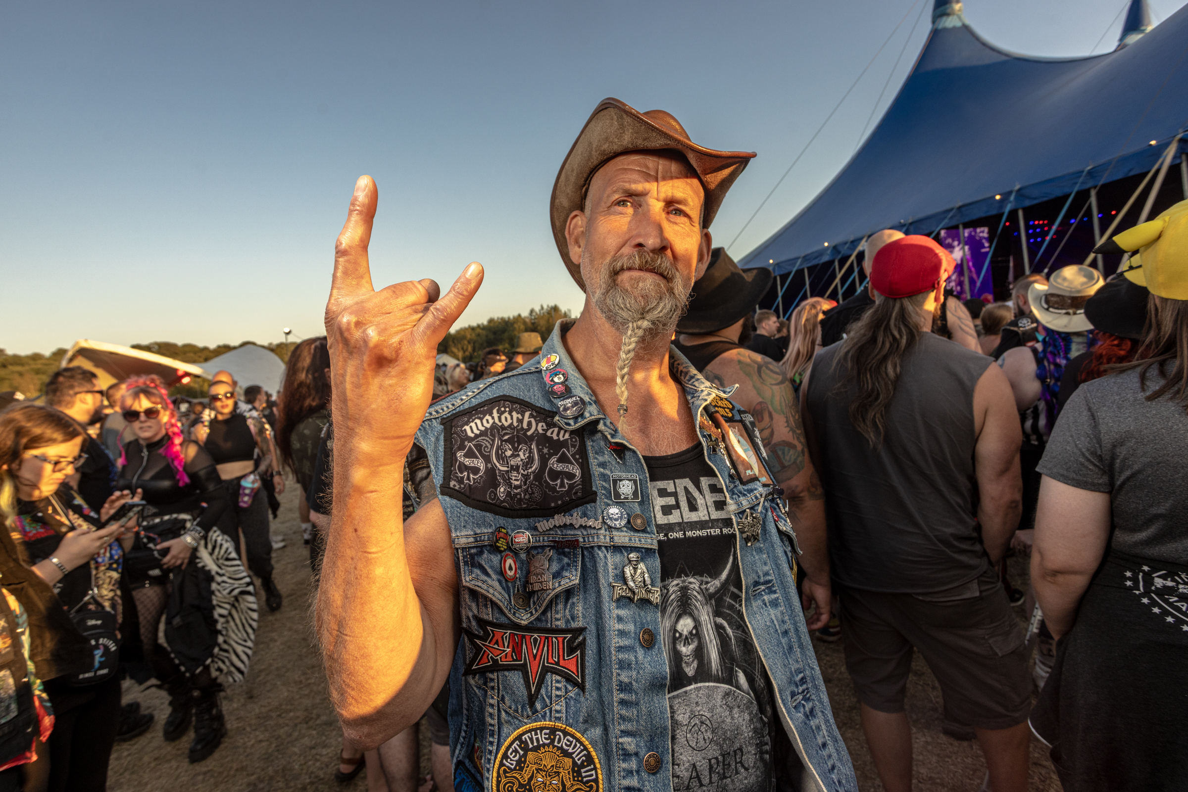 Bearded man in cowboy hat and patch-covered denim vest making rock horns gesture at outdoor festival with blue tent and crowd.