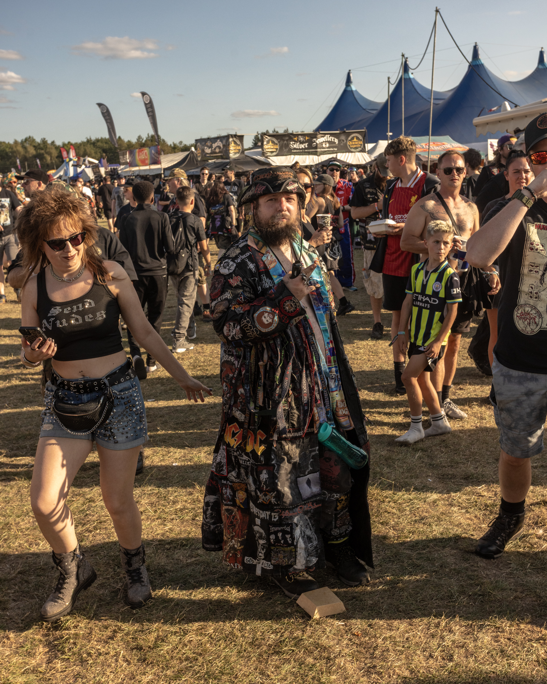 Crowd of people at outdoor music festival with blue peaked tents, food stalls, and banners in background under cloudy sky.