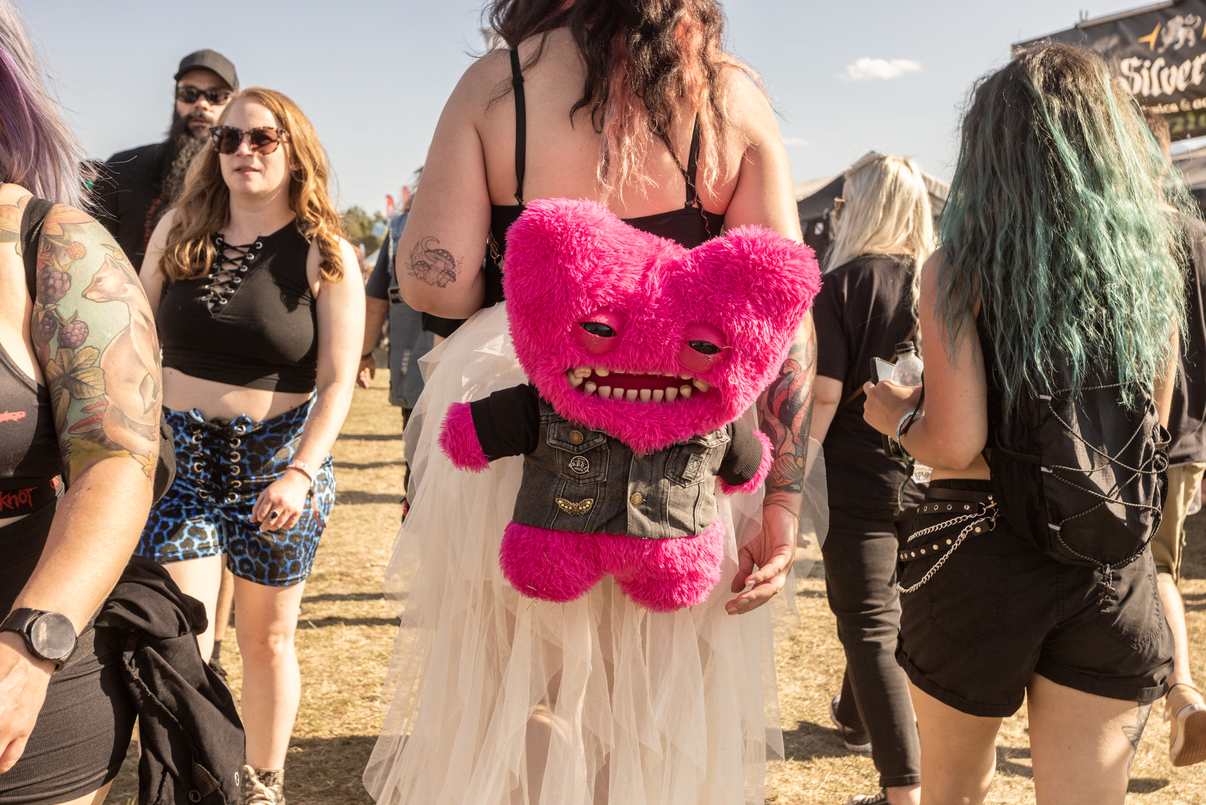 Woman in white dress holds bright pink furry monster toy with sharp teeth at outdoor festival with other attendees in black clothing.