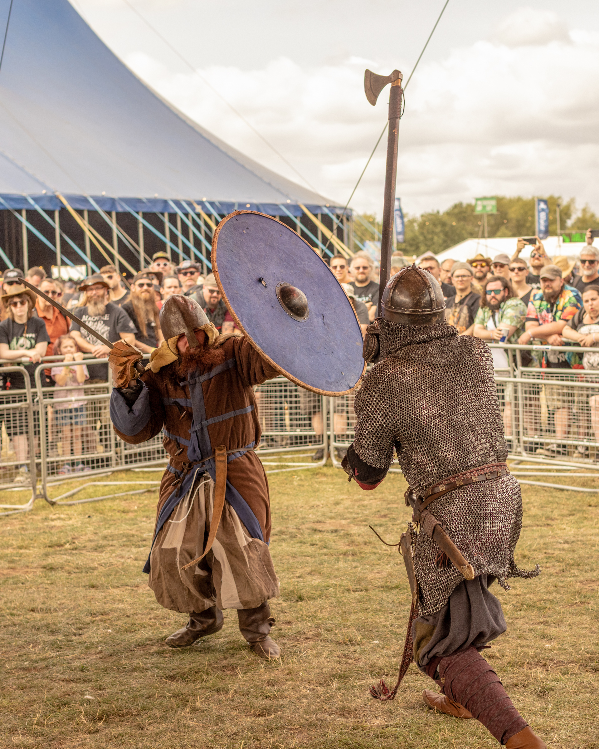 Two people in medieval costume engaged in combat reenactment, one holding shield and axe, other in chainmail with sword, crowd watching.