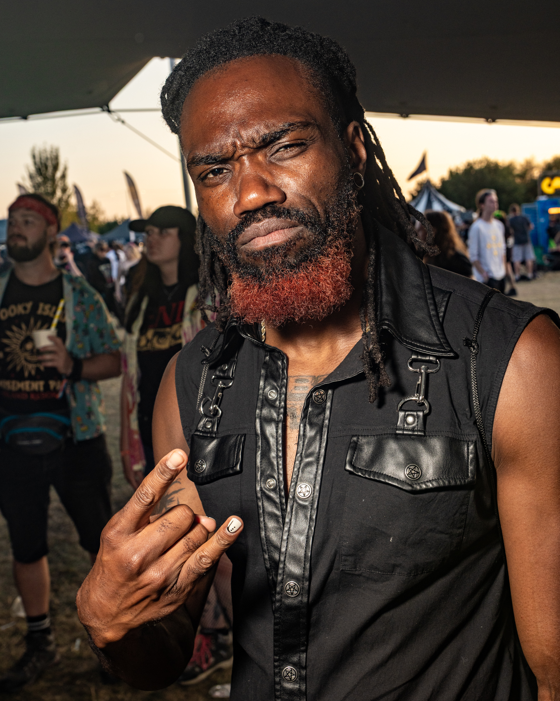 Black man with long dreadlocks and beard wearing black leather waistcoat at outdoor event with crowd in background.