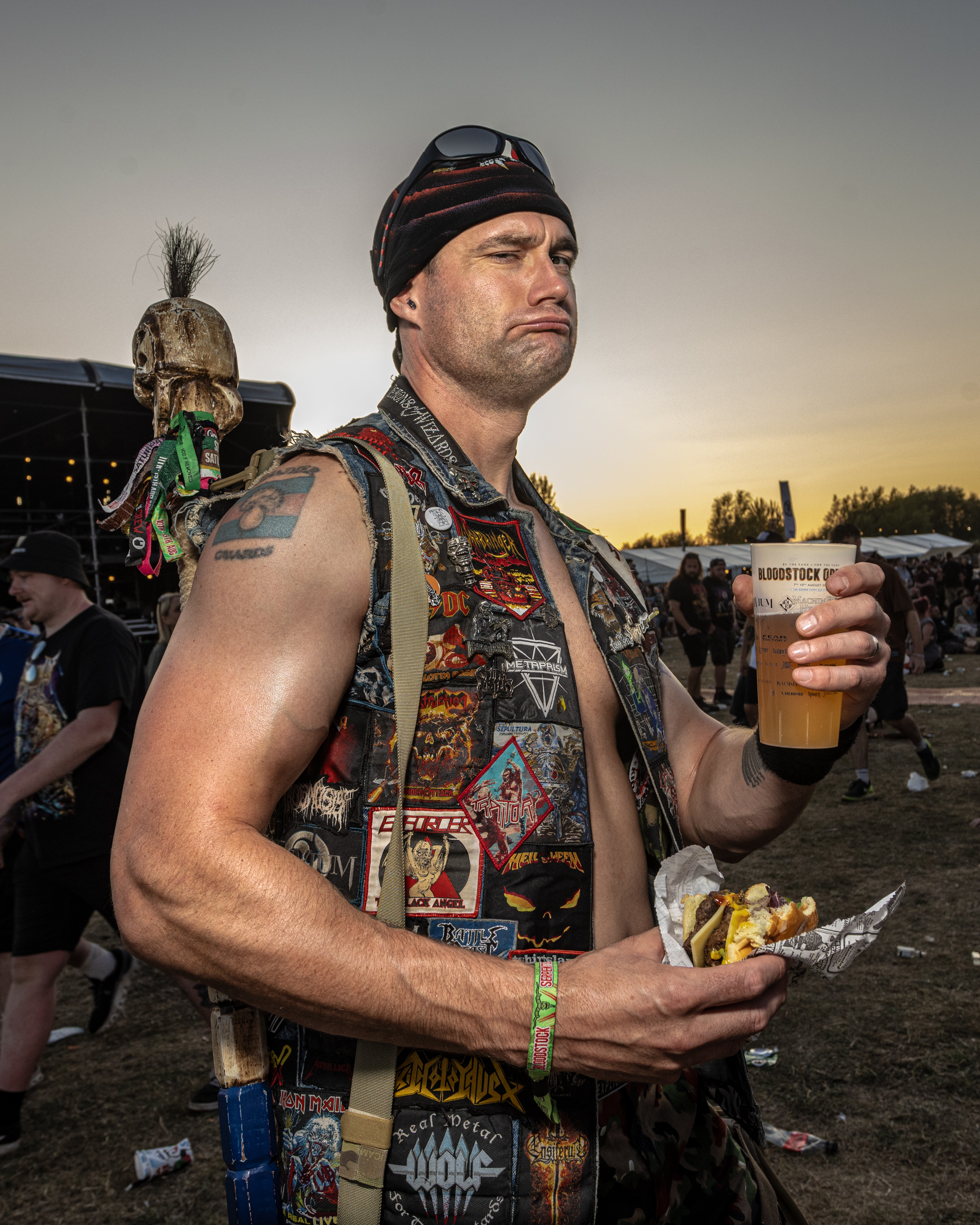 Man in black beanie and patch-covered vest holding pint and food at outdoor festival, with decorative skull visible in background.