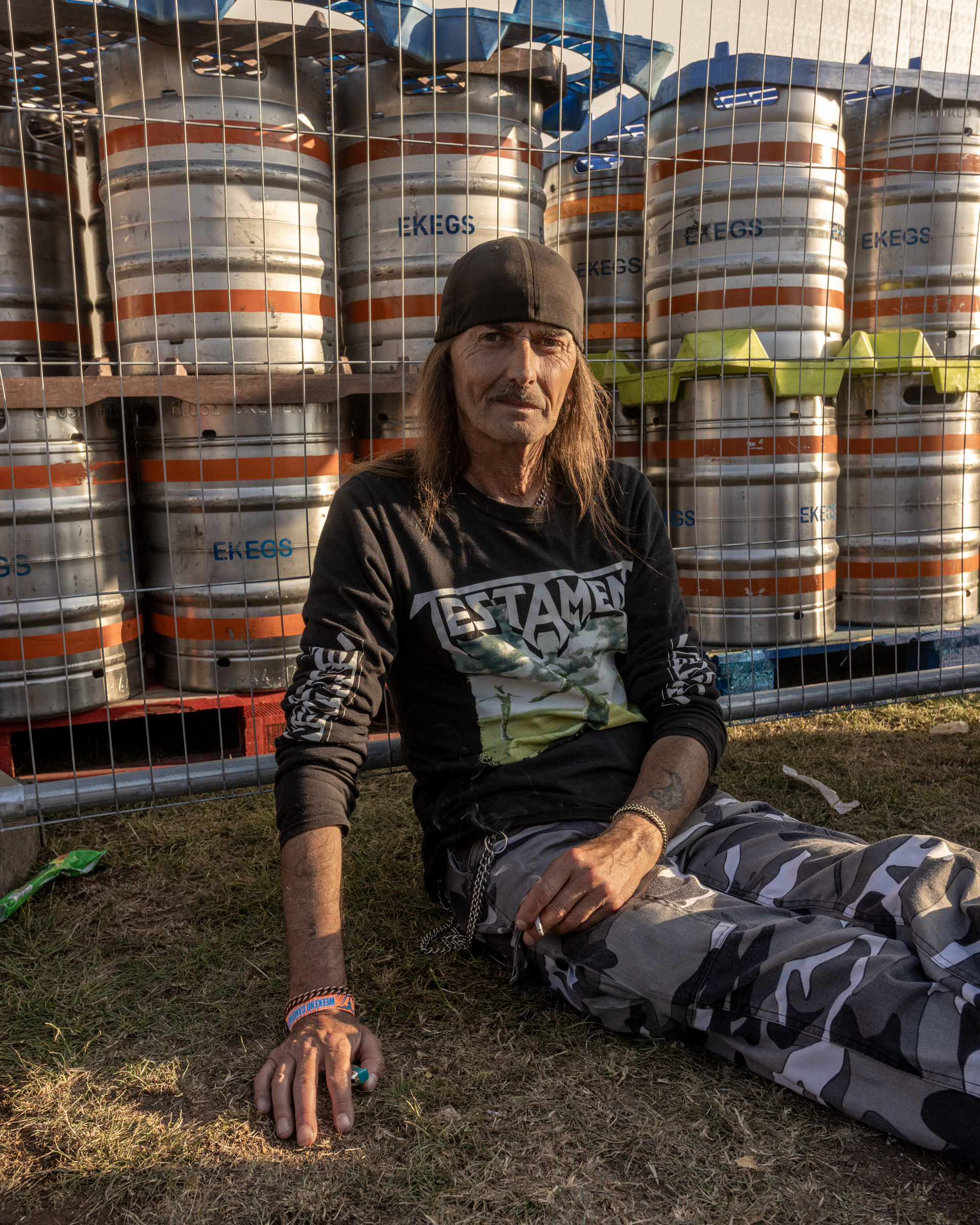 Man with long hair and beanie sits on ground wearing dark top, with stacked metal kegs behind wire fencing in background.