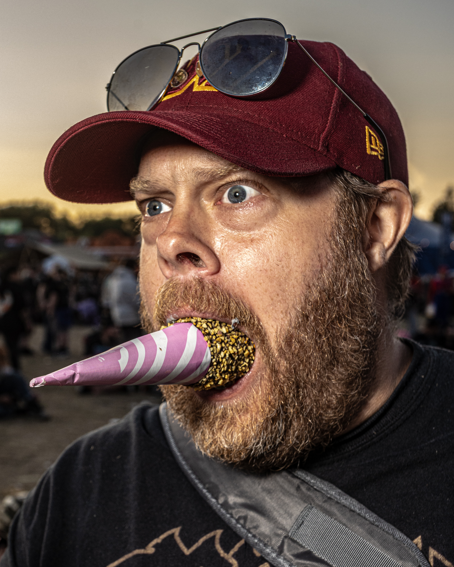 Bearded man wearing burgundy baseball cap and sunglasses with pink and white striped party horn in mouth covered in gold glitter.