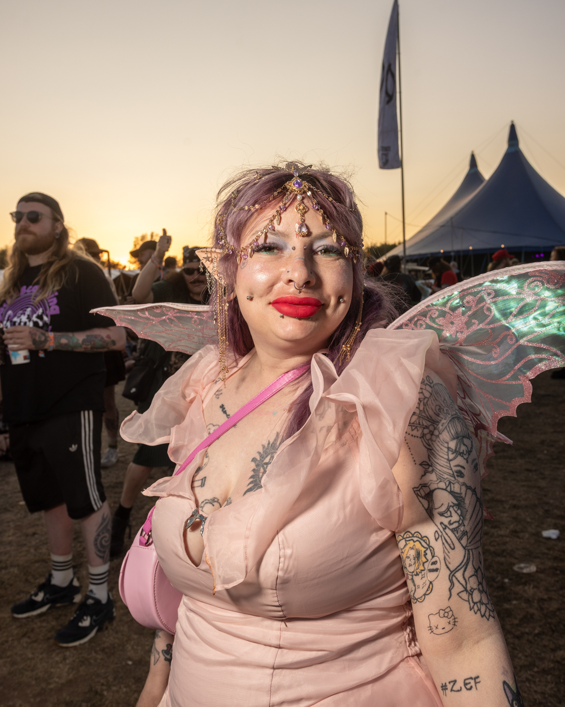 Person in pink fairy costume with wings, gold head chain, and red lipstick at outdoor festival with blue tents and crowd in background.