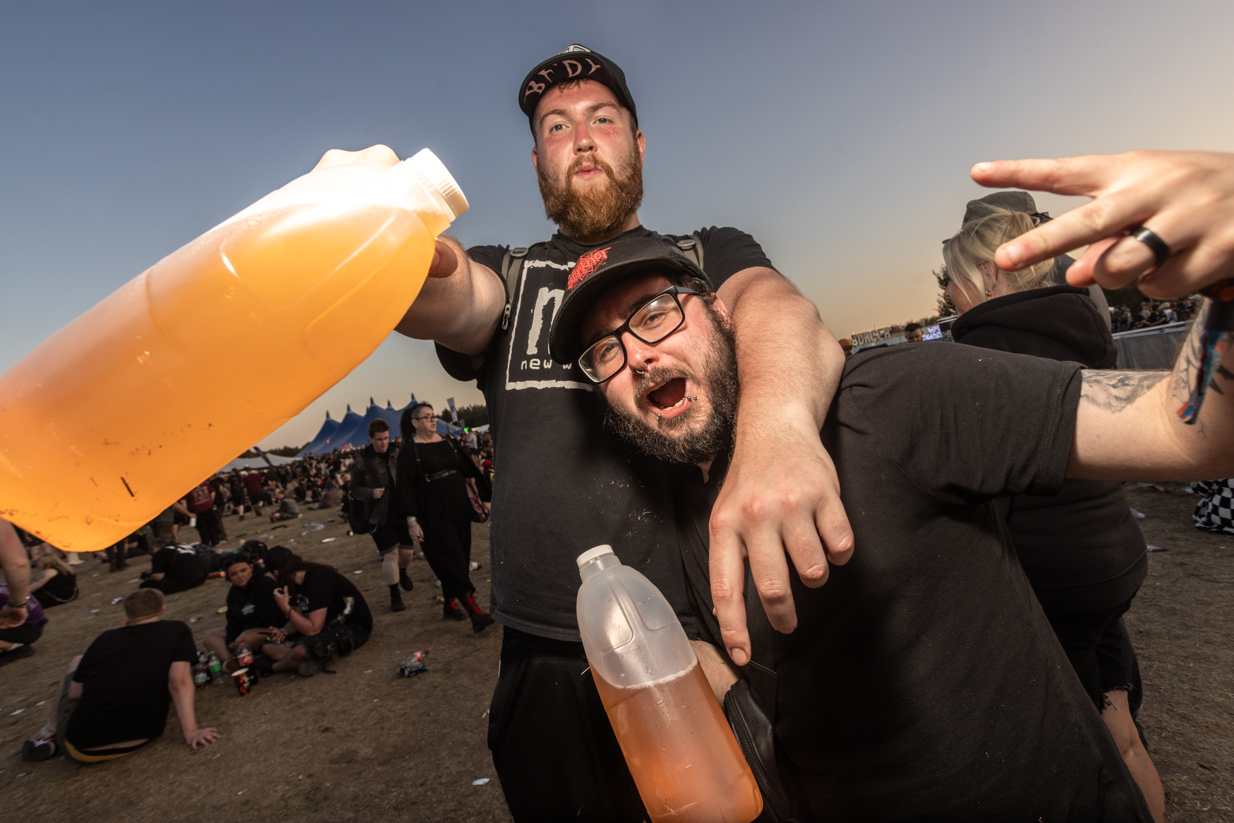 Two bearded men holding plastic cups of orange beer at outdoor event with crowd and blue-orange sunset sky in background.