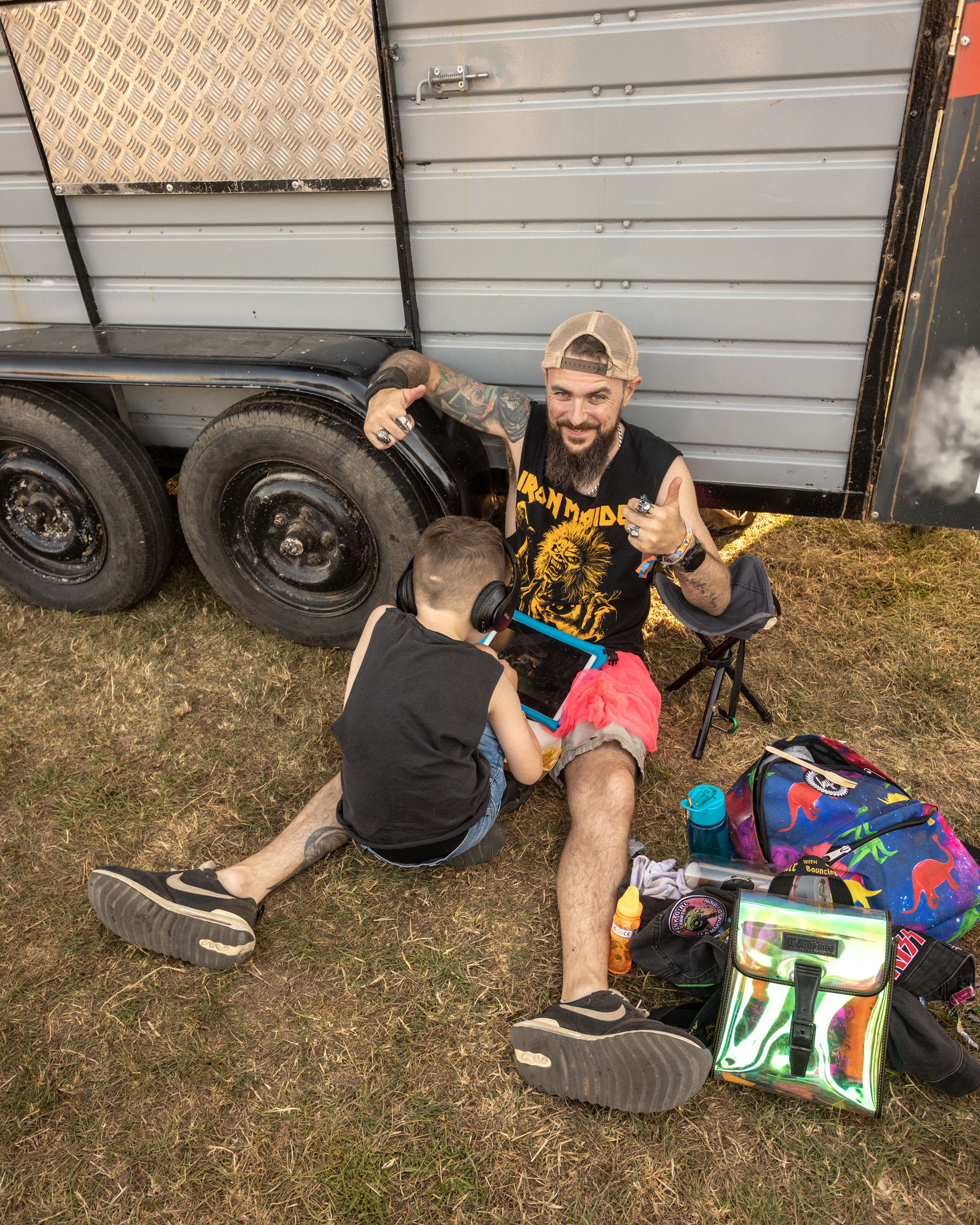 Bearded man in cap getting tattoo from artist beside grey trailer, camping chairs and colourful bags on grass ground.