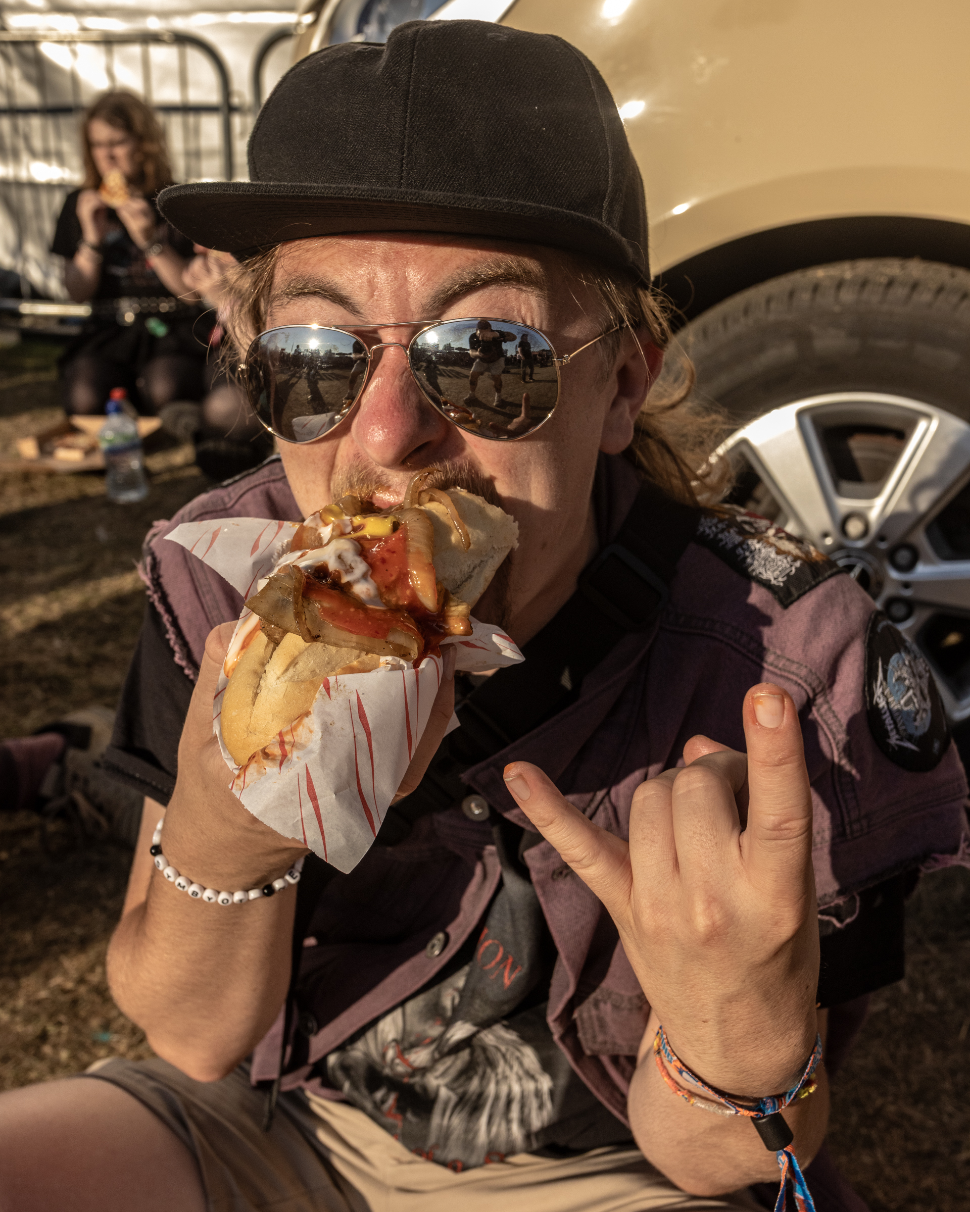 Man in black cap and sunglasses eating sandwich whilst making peace sign gesture, with woman and car visible in background