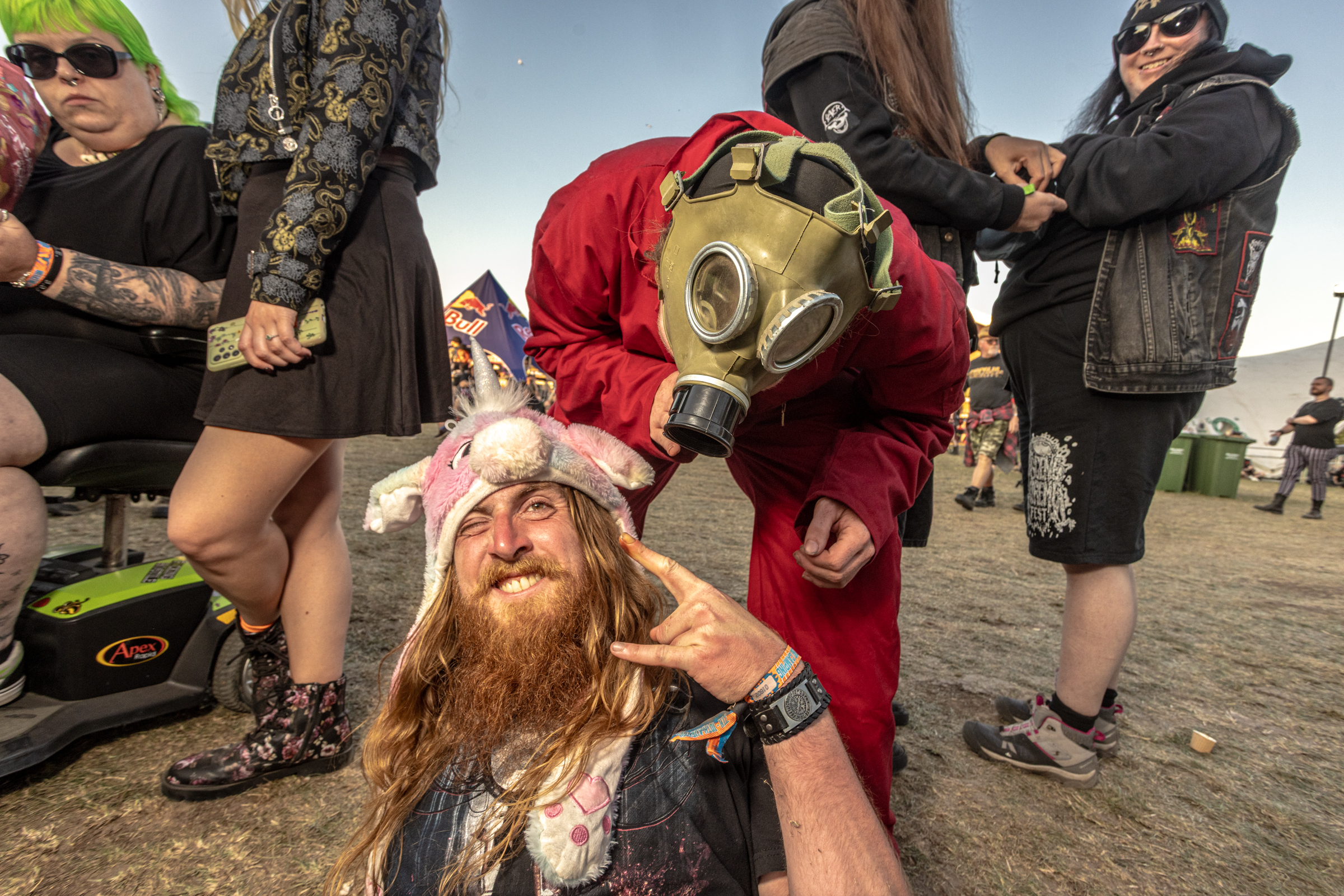 Person in gas mask and red jacket crouched behind bearded man with long hair making peace sign, surrounded by people in sunglasses outdoors.
