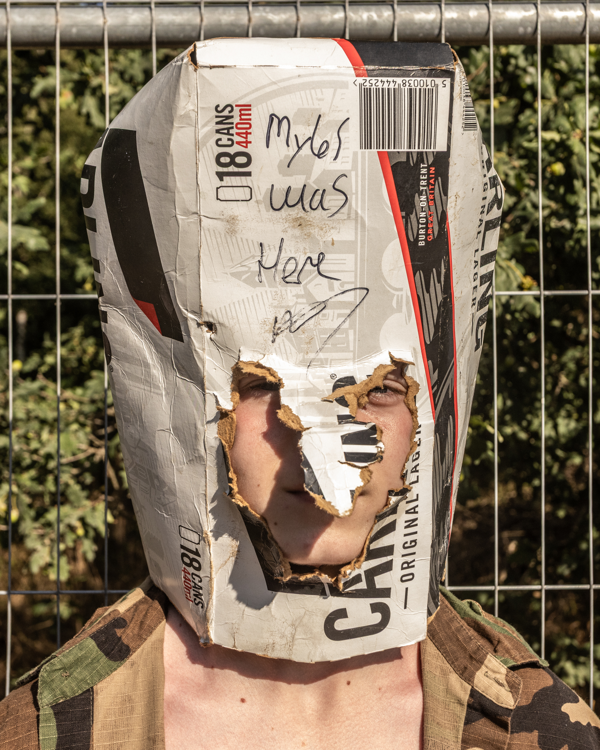 Person wearing torn white paper bag over head with "Myles was here" handwritten in black text, standing against wire fence.