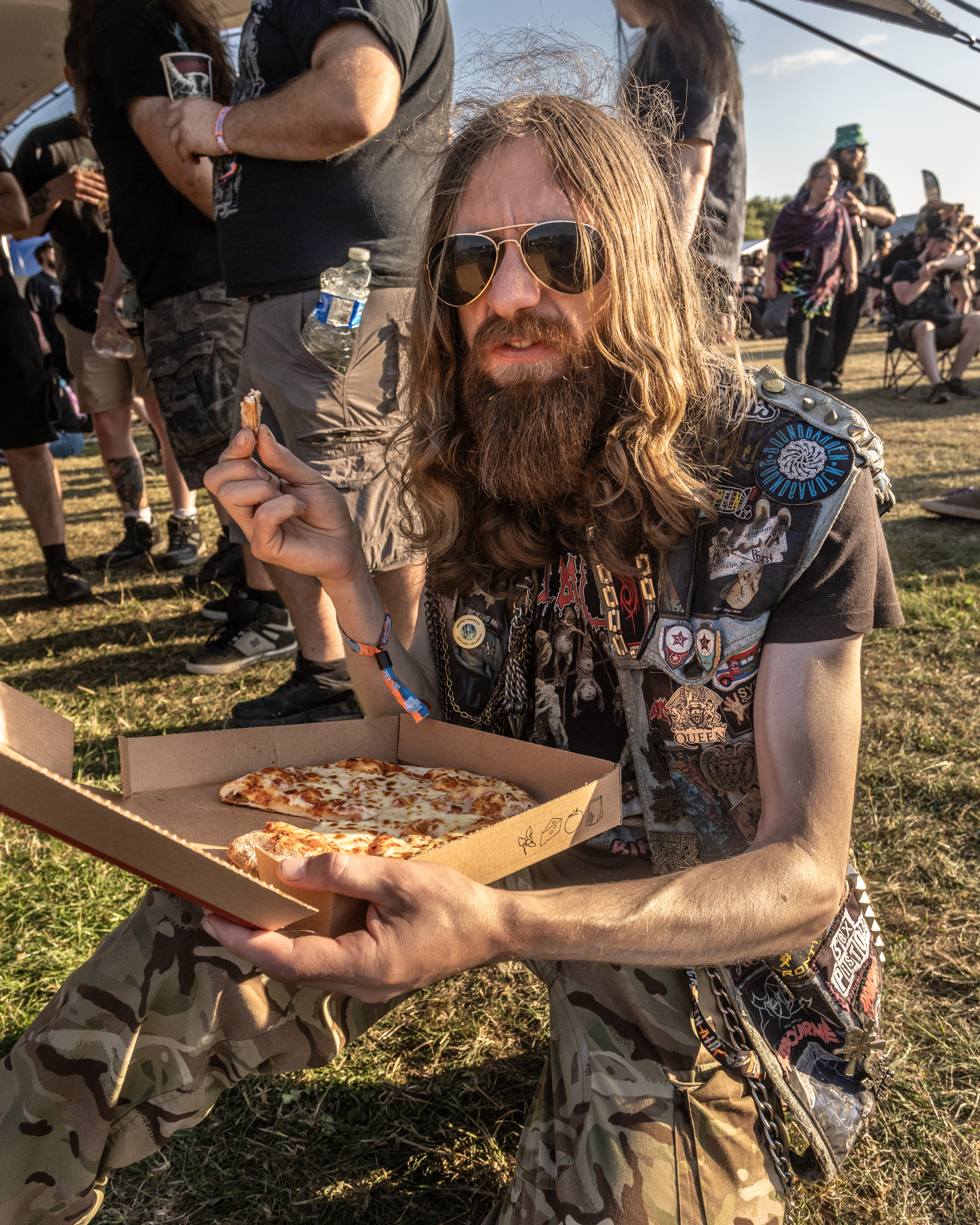 Long-haired bearded man in sunglasses and patch-covered vest eating pizza slice from box at outdoor festival with crowd behind him.