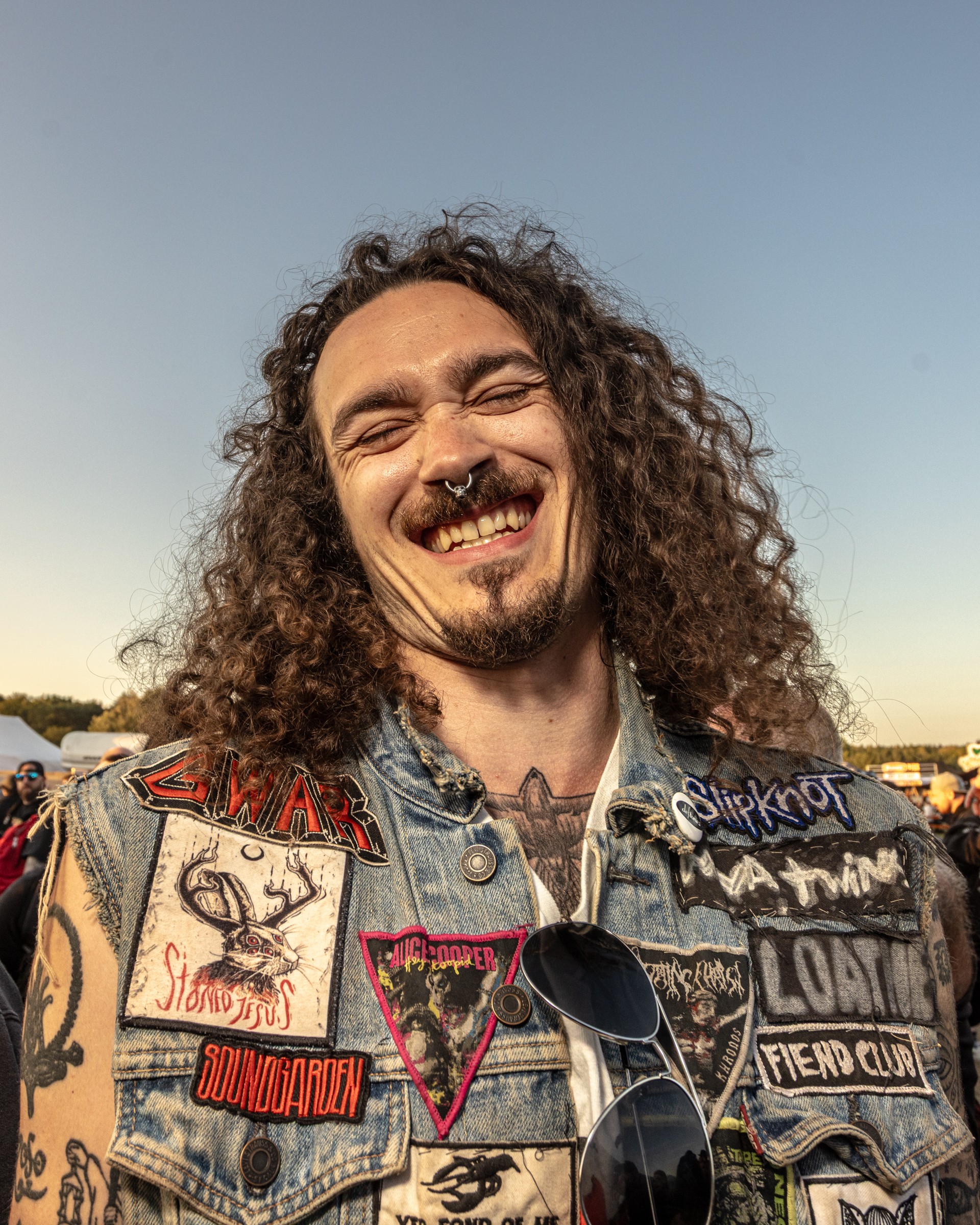 Man with long curly dark hair and beard smiling, wearing denim jacket covered in metal band patches against blue sky.