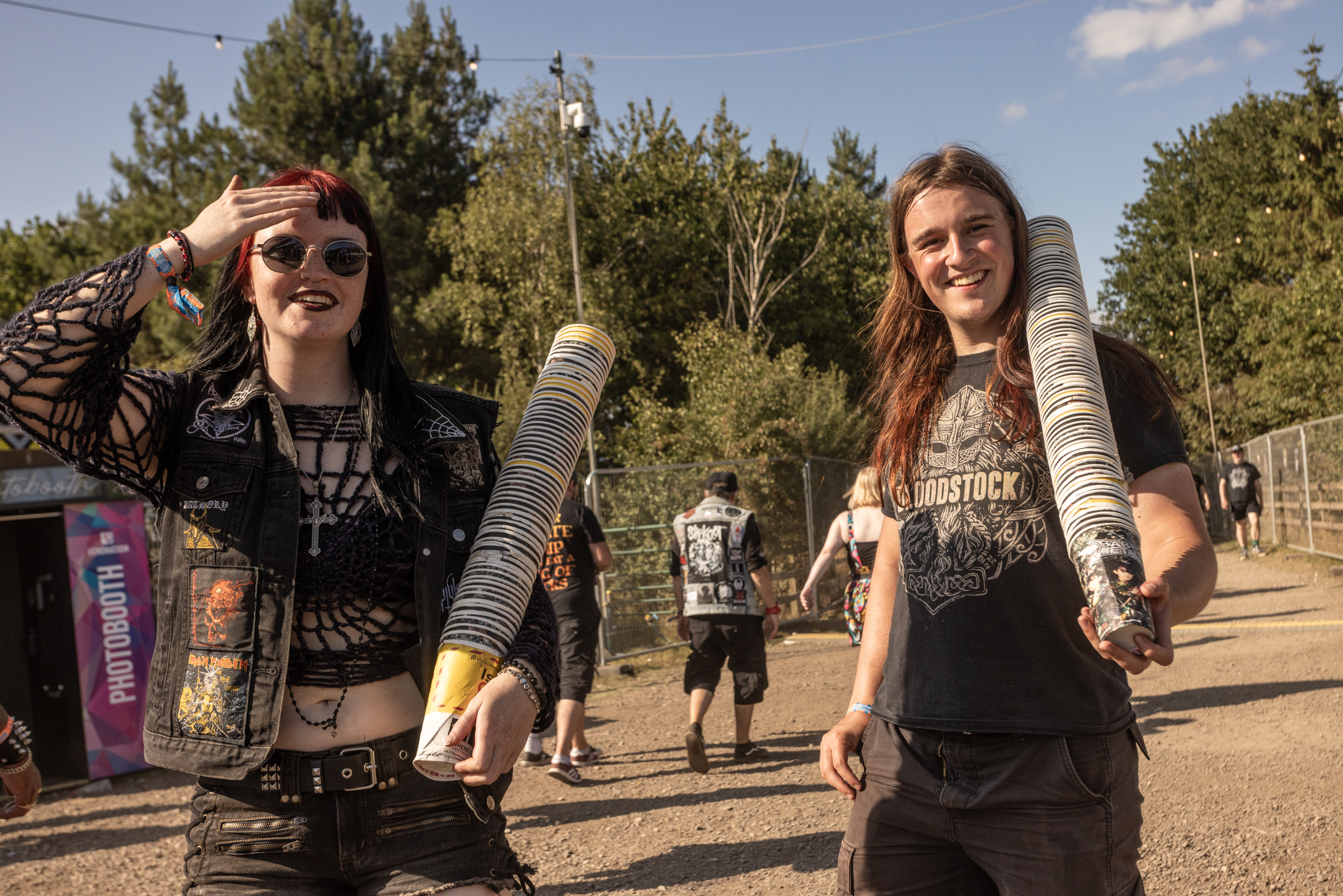 Two women standing outdoors holding skateboards, wearing casual clothing and sunglasses, with trees and people in background.