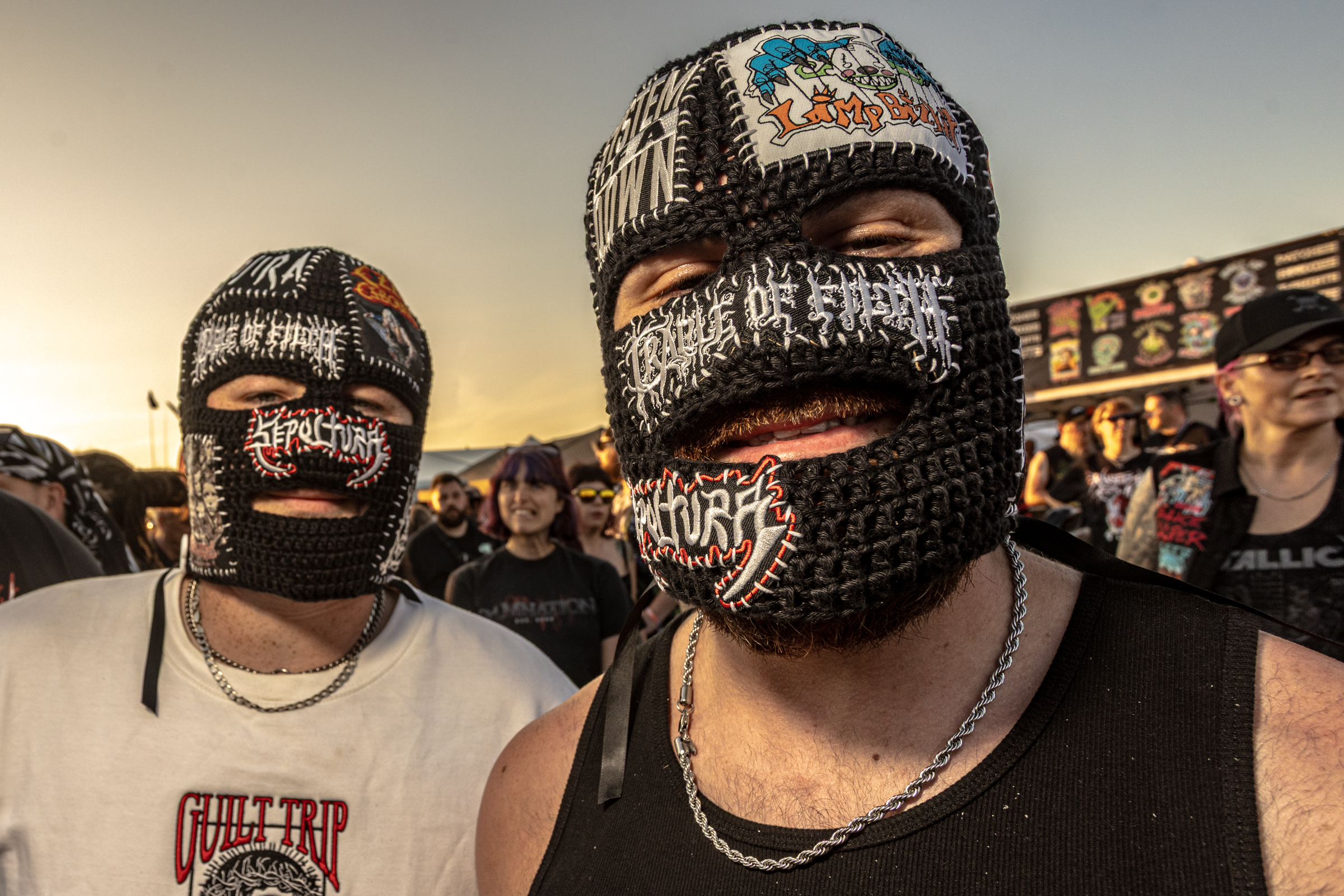 Two men wearing knitted balaclavas with colourful patterns and text embroidery, standing in crowd at outdoor event during sunset.