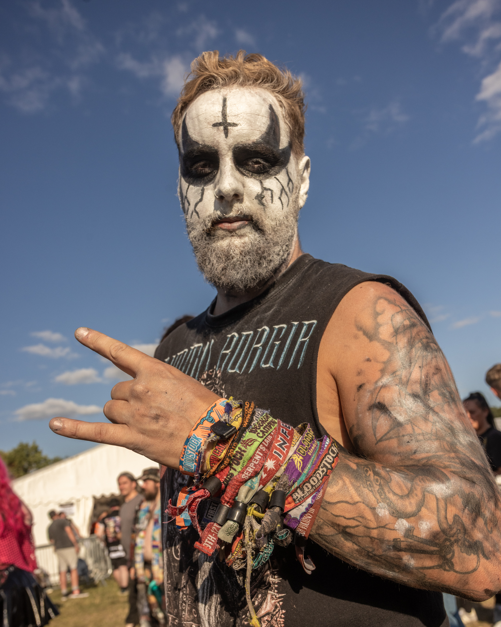 Bearded man with black and white face paint featuring cross and teardrop designs, wearing band t-shirt, making hand gesture outdoors.