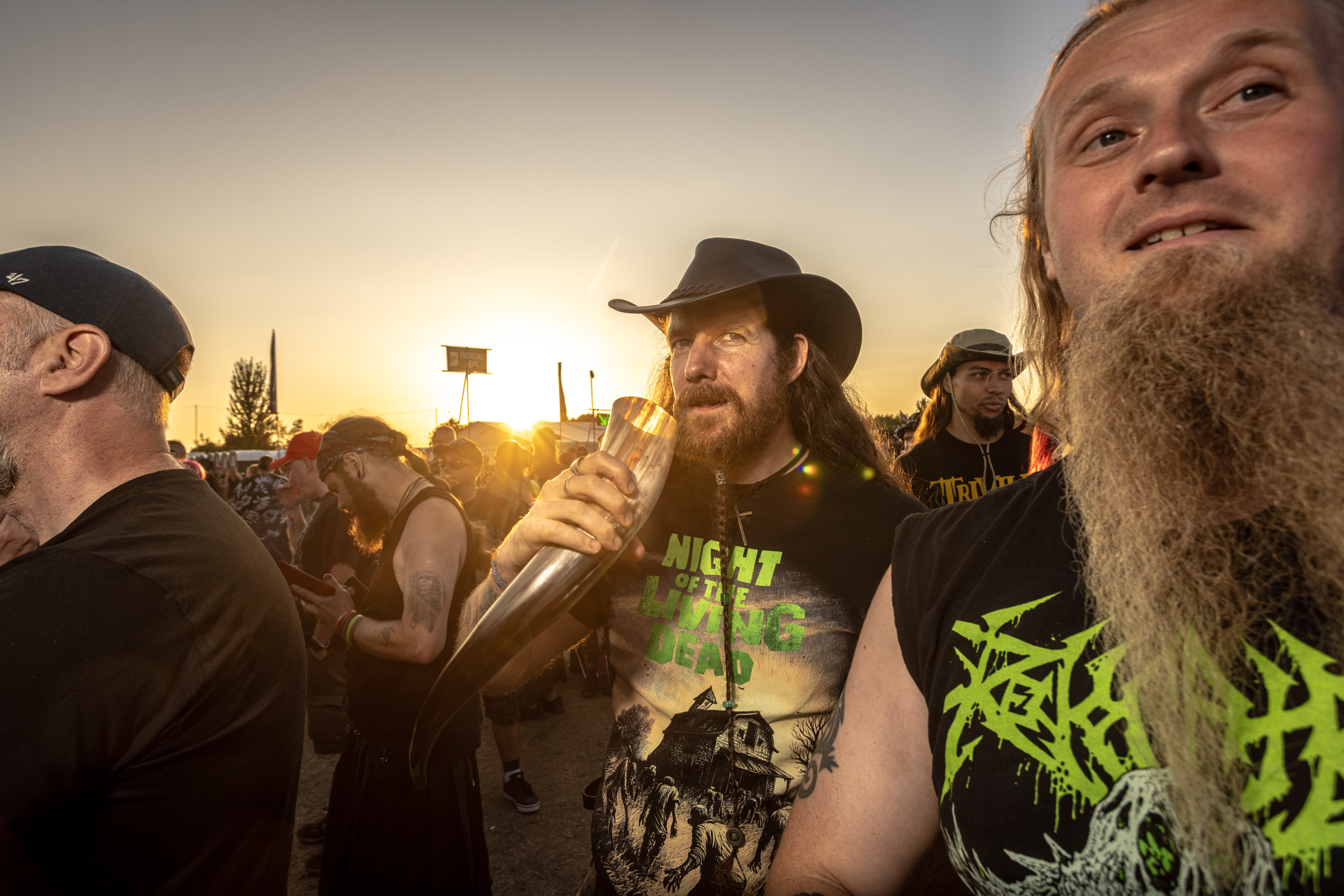 Bearded man in black hat holding microphone at outdoor gathering, crowd visible in background, sunset lighting.