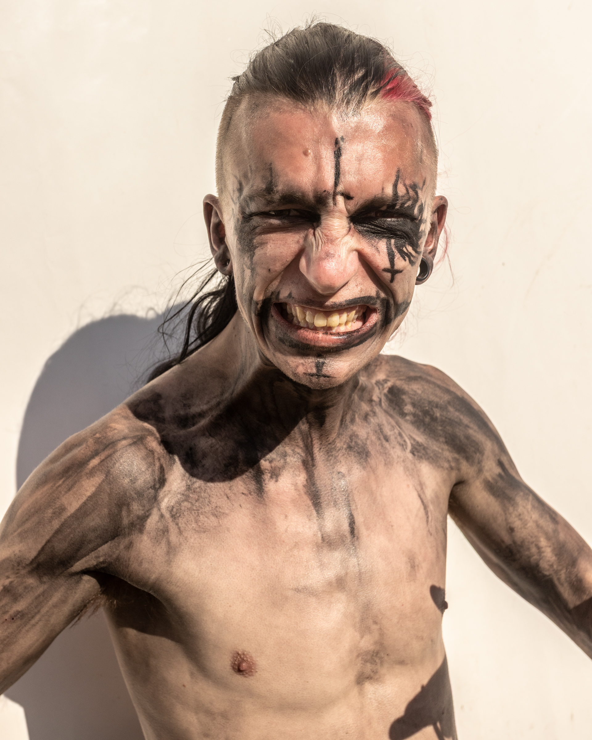 Shirtless man with dark tribal-style face paint and body markings, grinning widely with arms raised against light background.