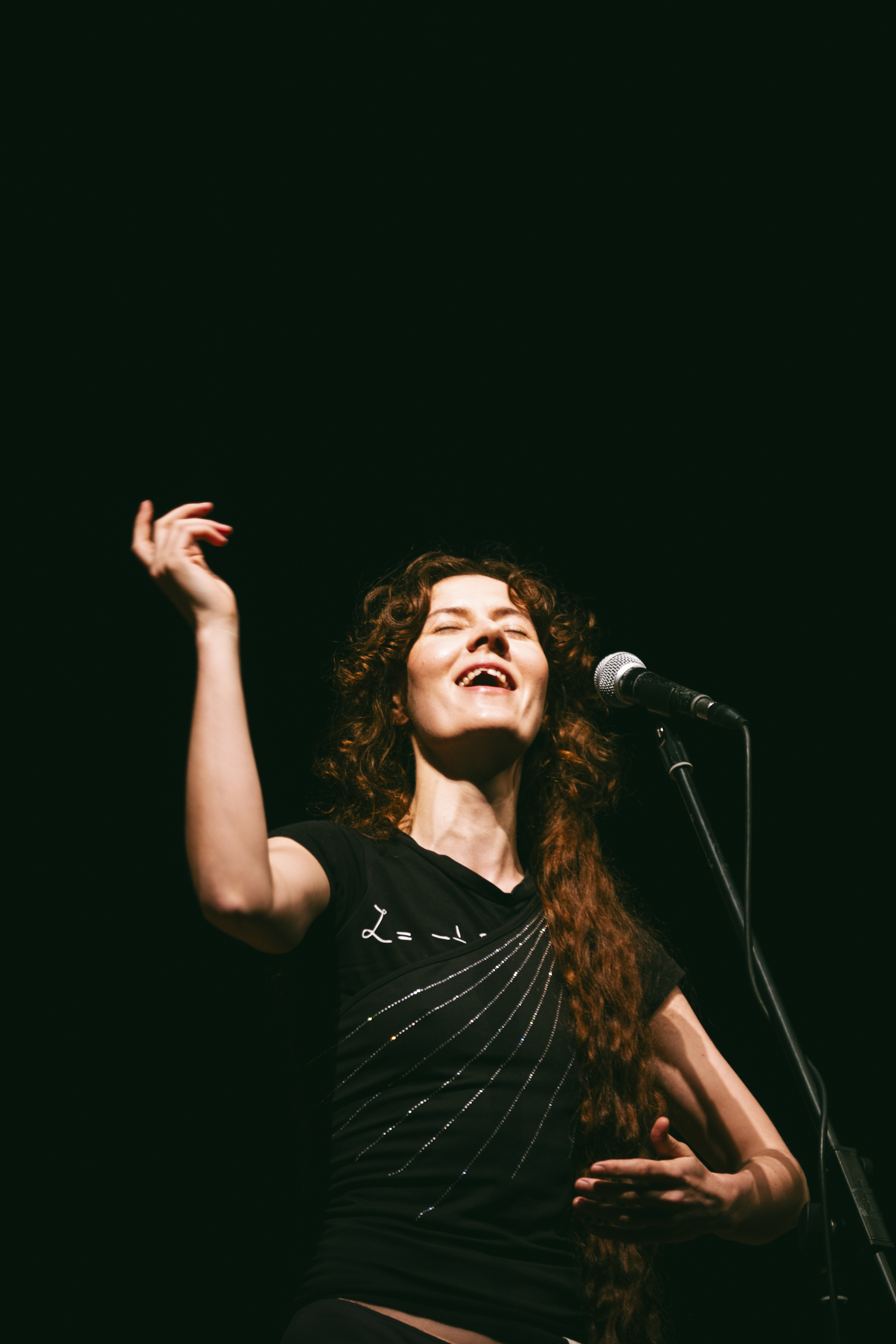 A woman with curly hair singing into a microphone on stage, wearing a black shirt and raising one arm.
