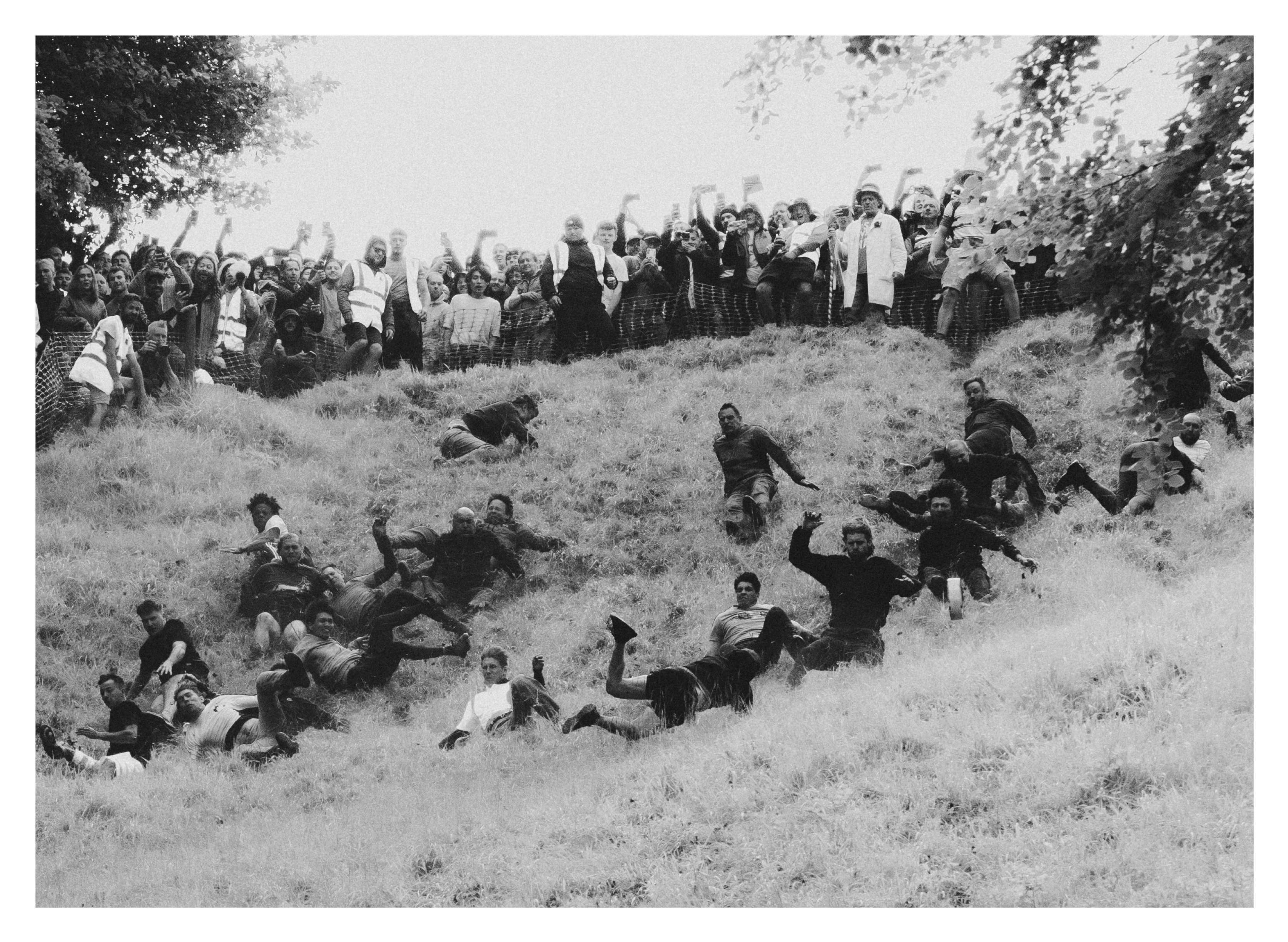 Black and white image showing people tumbling down a steep grassy hill whilst crowd watches from top edge, trees visible on sides.