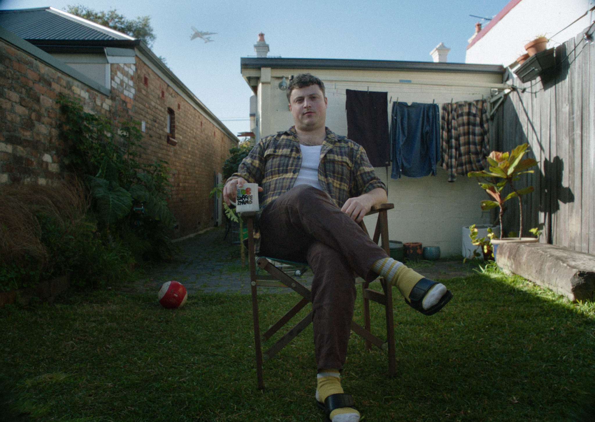 Man sits on wooden chair in garden with brick walls, white building, and washing line with clothes hanging in background.