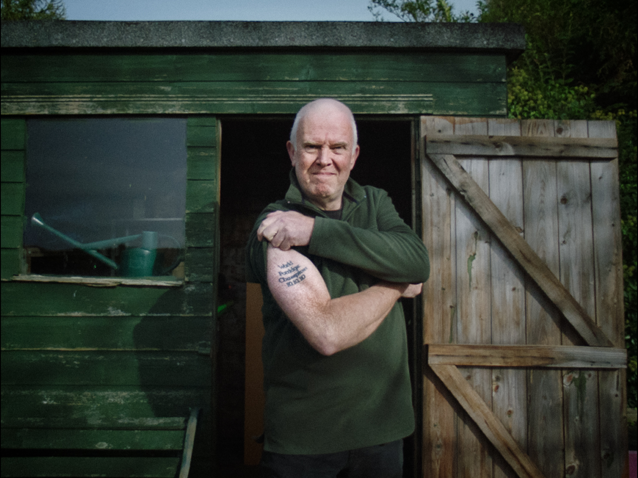 Elderly man with white beard in green t-shirt standing by weathered wooden shed, arms crossed, showing tattoo on forearm.