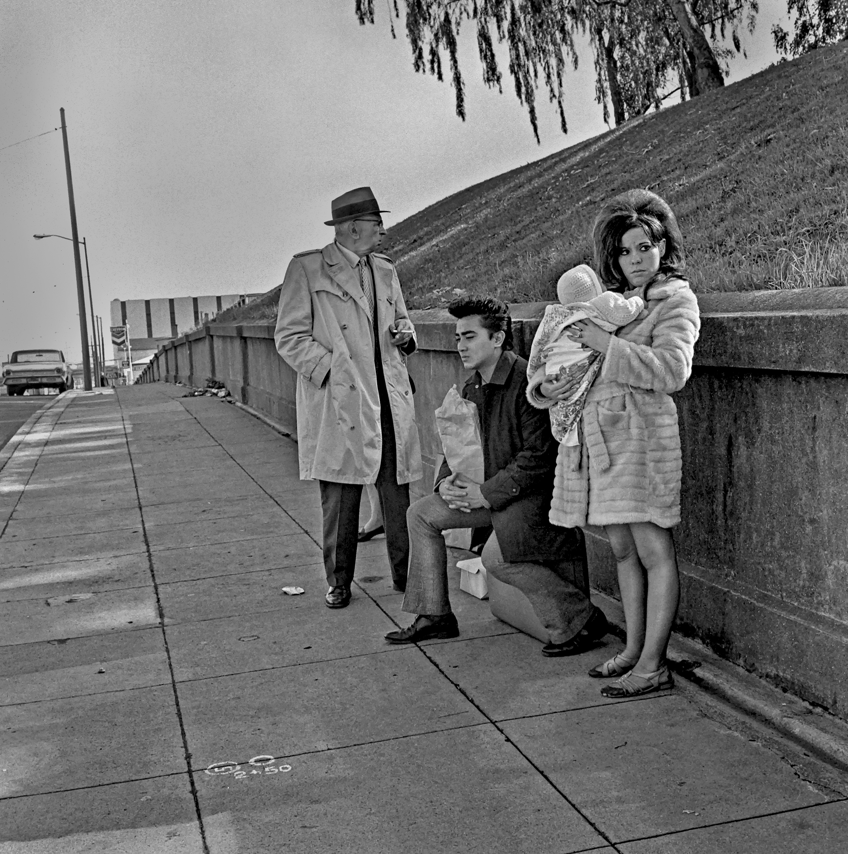 A group of people standing on a sidewalk near a building. The image is in black and white and shows a man in a hat, a seated person, and a woman holding a child.