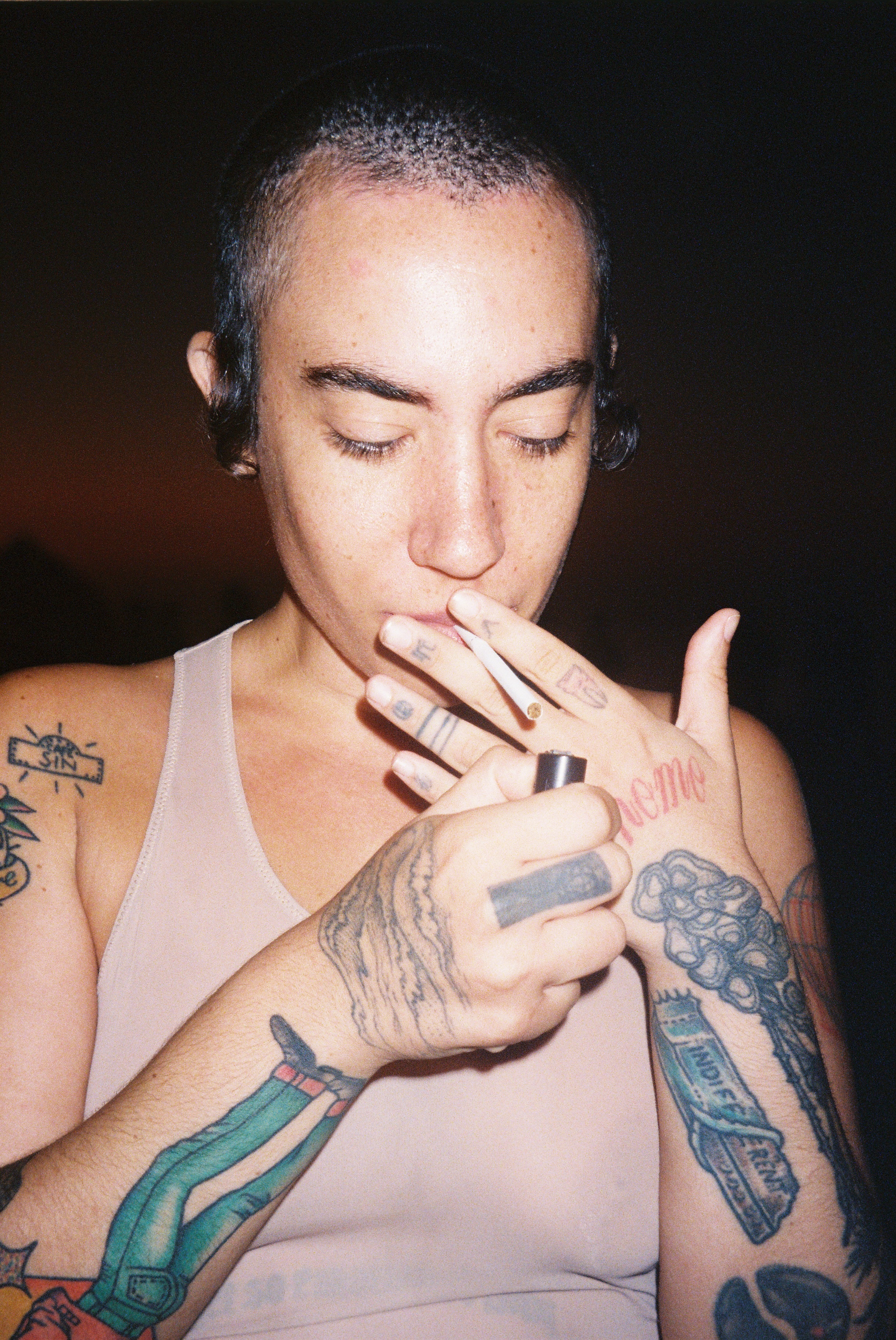A tattooed individual with their eyes closed, holding a can or spray bottle near their mouth in a dark setting.