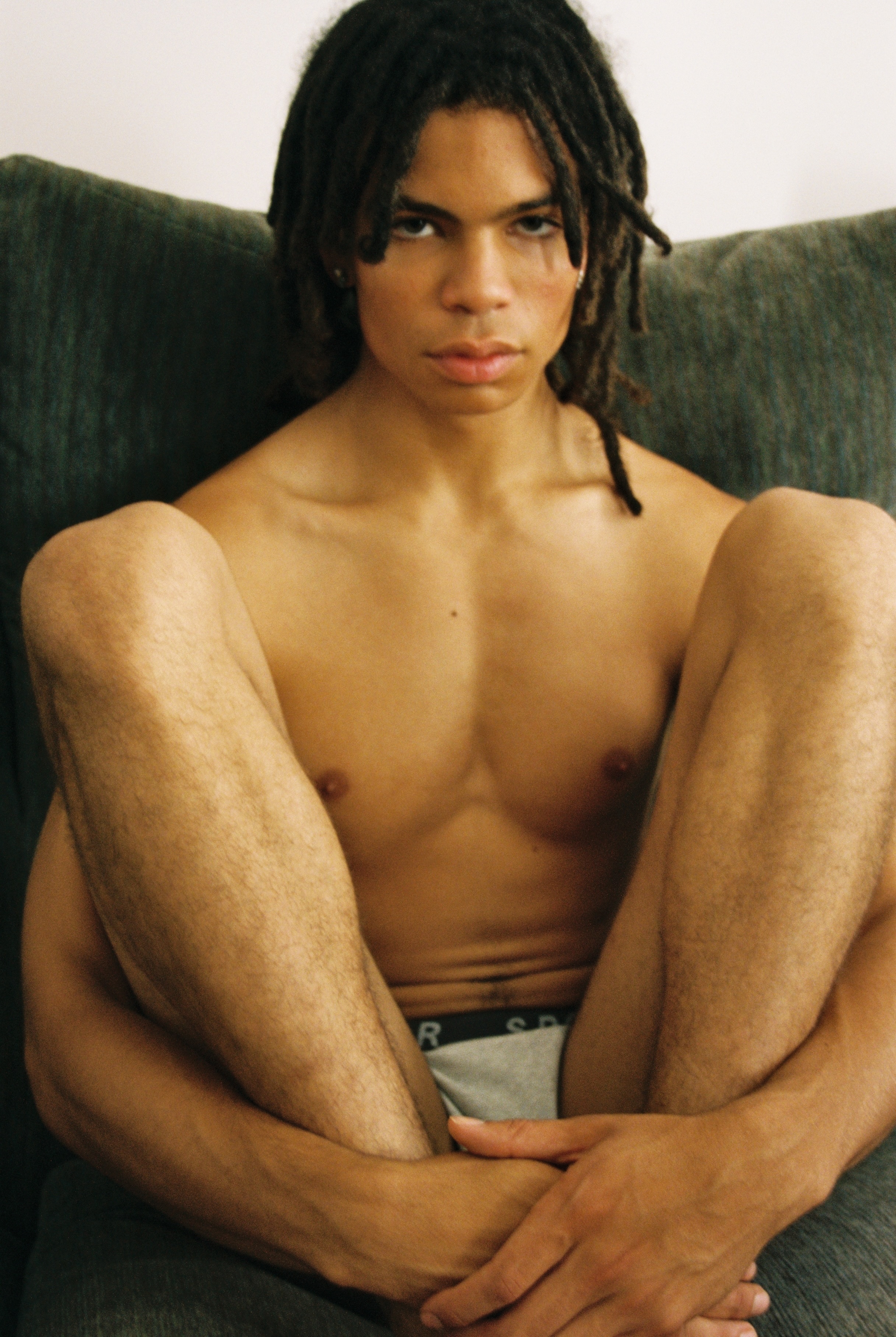 Image of a shirtless man with dreadlocks, sitting on a sofa and looking at the camera.