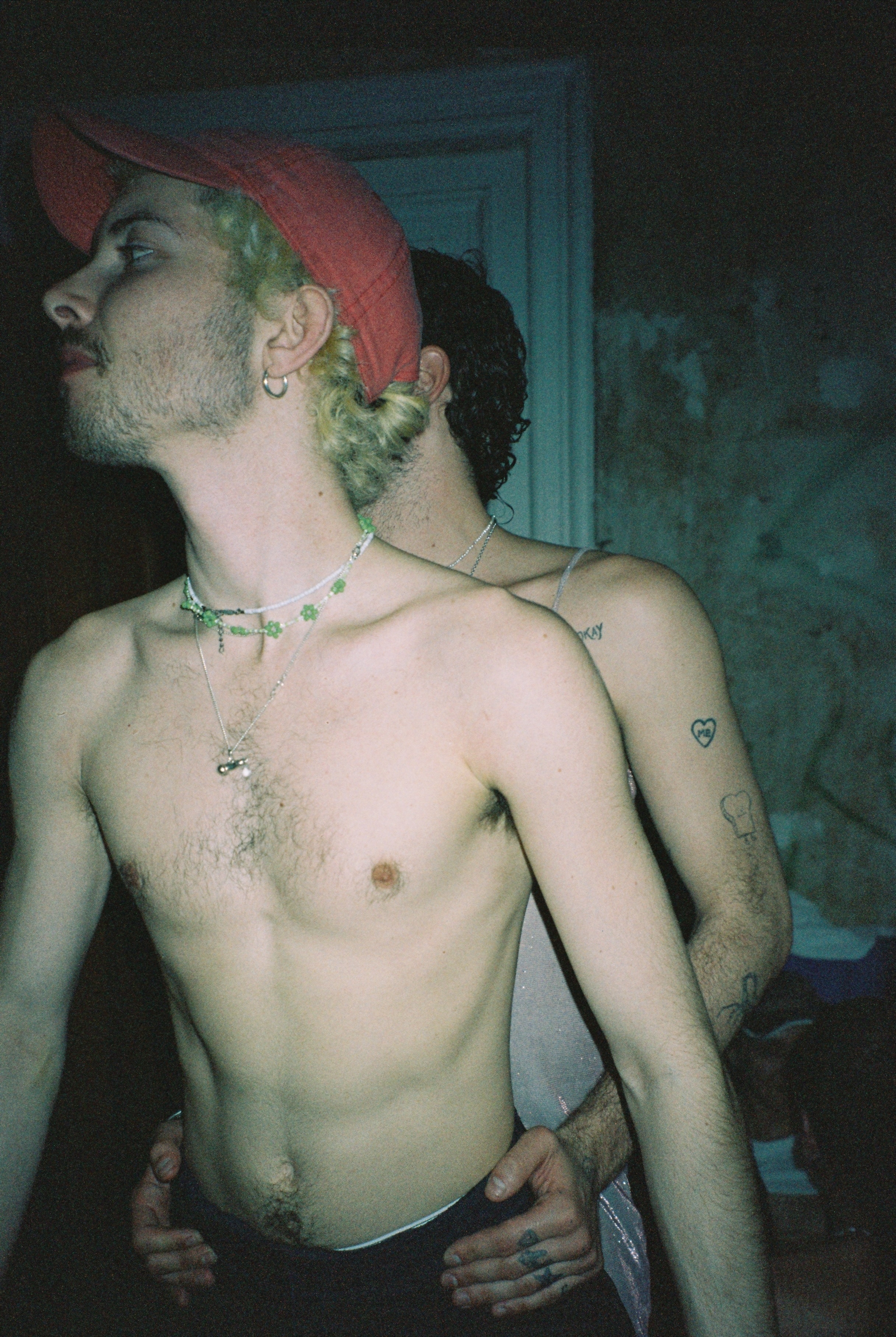 A person with green hair and tattoos, wearing a red hat, shown in a shadowy setting.